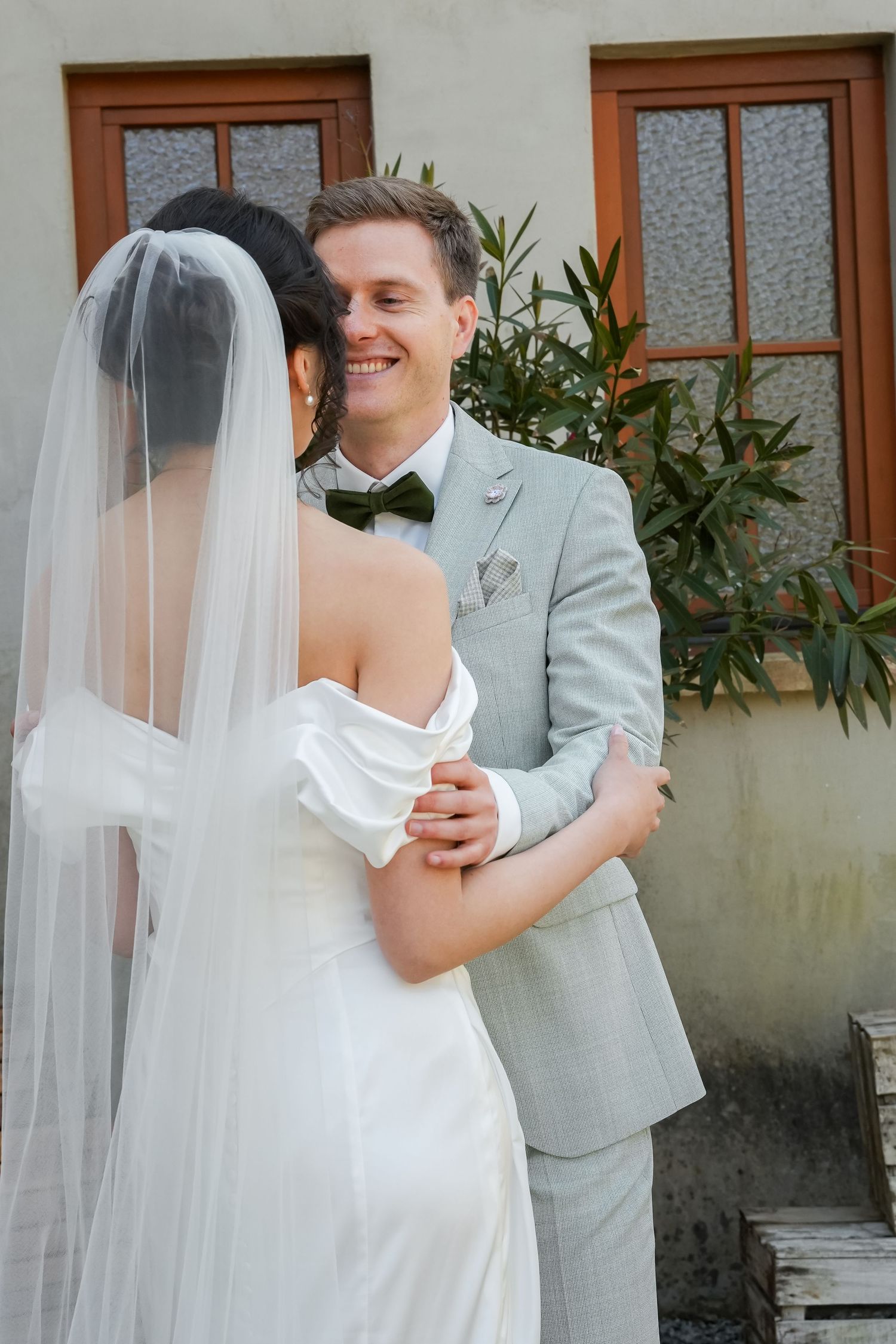 Smiling groom in gray suit embraces bride in white off-shoulder gown and veil outdoors near a building.