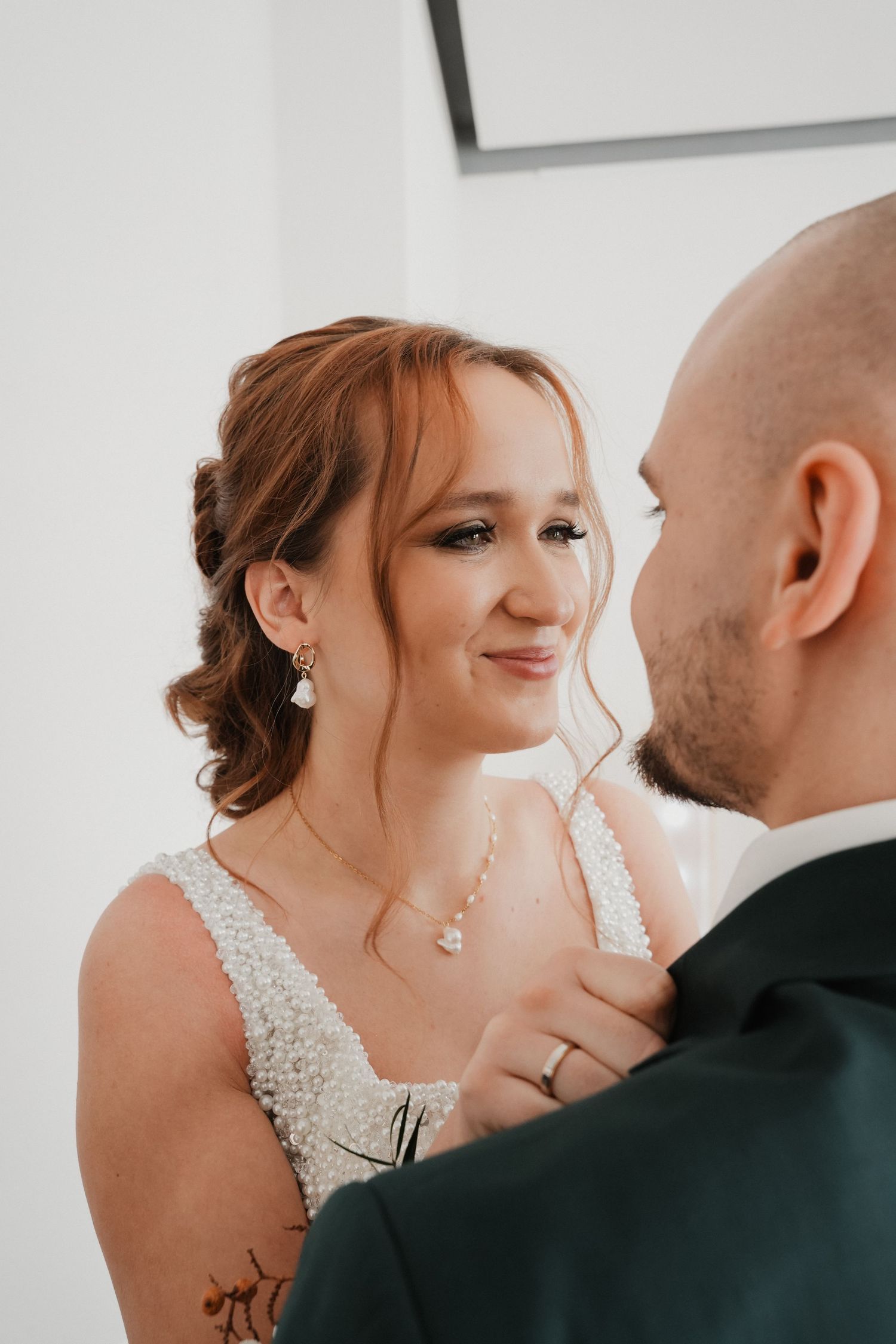 Bride in pearl wedding dress gazes lovingly at groom in green suit during intimate wedding moment.