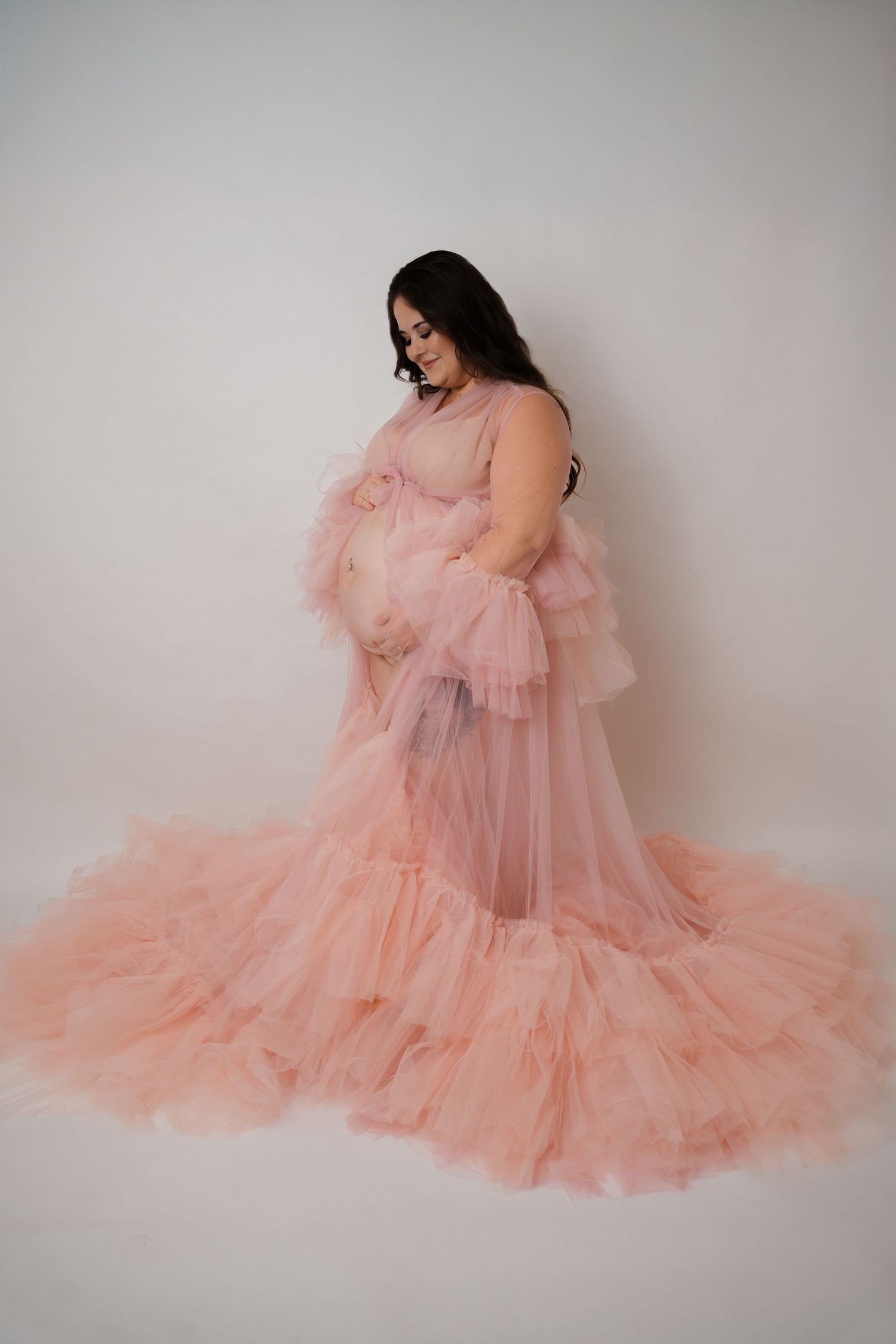 Pregnant woman in a flowing pink tulle maternity gown poses gracefully against a white studio background.