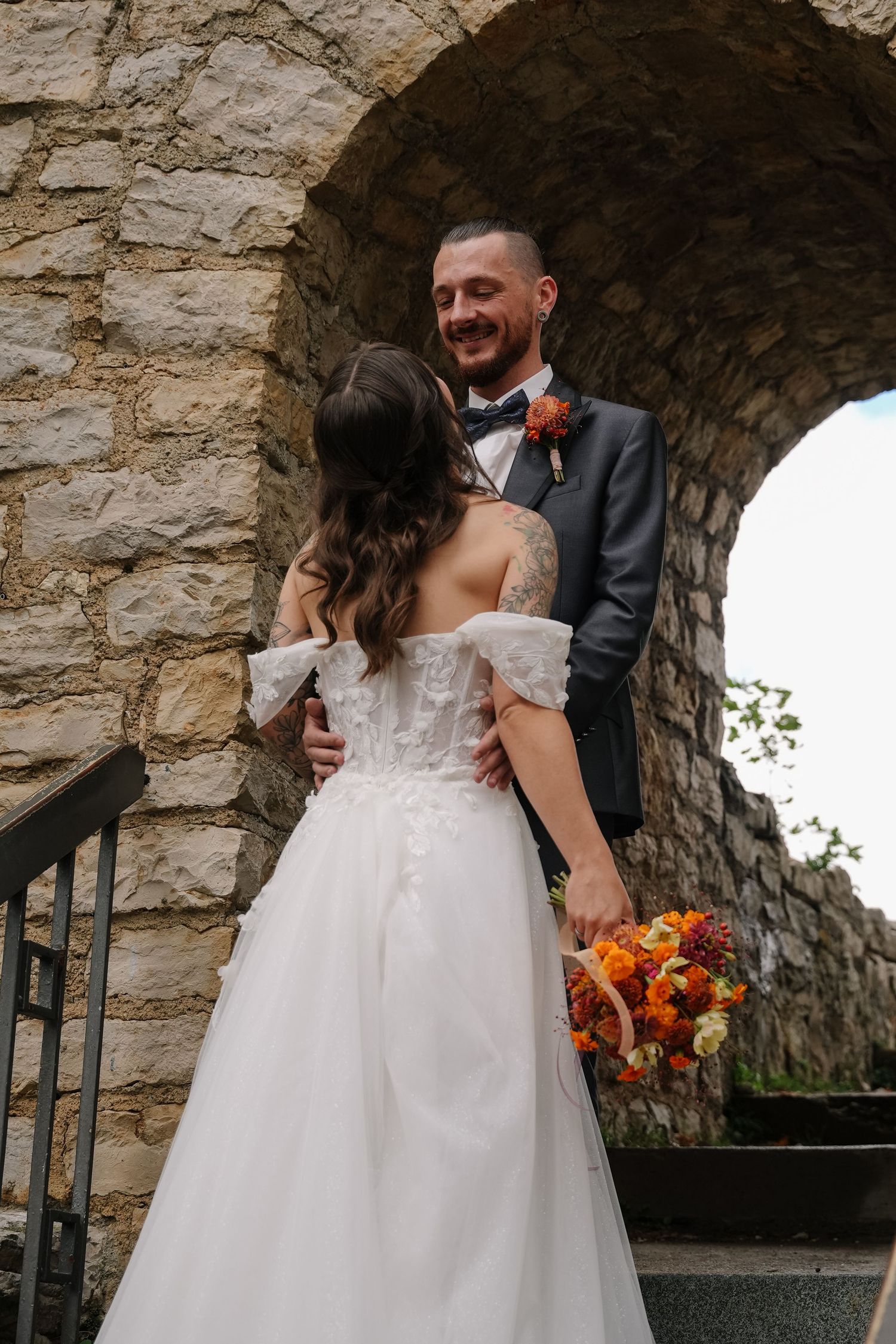 Bride in an off-shoulder tulle wedding gown holding an orange bouquet embraces her smiling groom beneath a rustic stone archway.