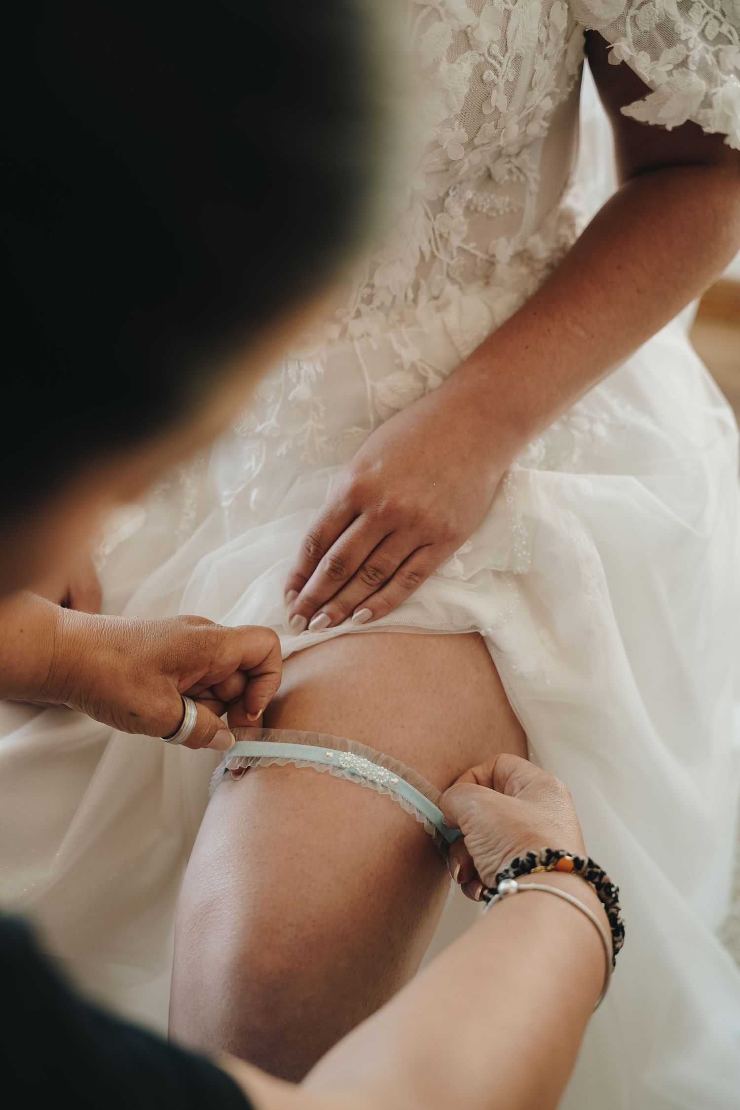 Bride in a white lace wedding dress has a light blue garter slipped onto her thigh by a helper wearing beaded bracelets.