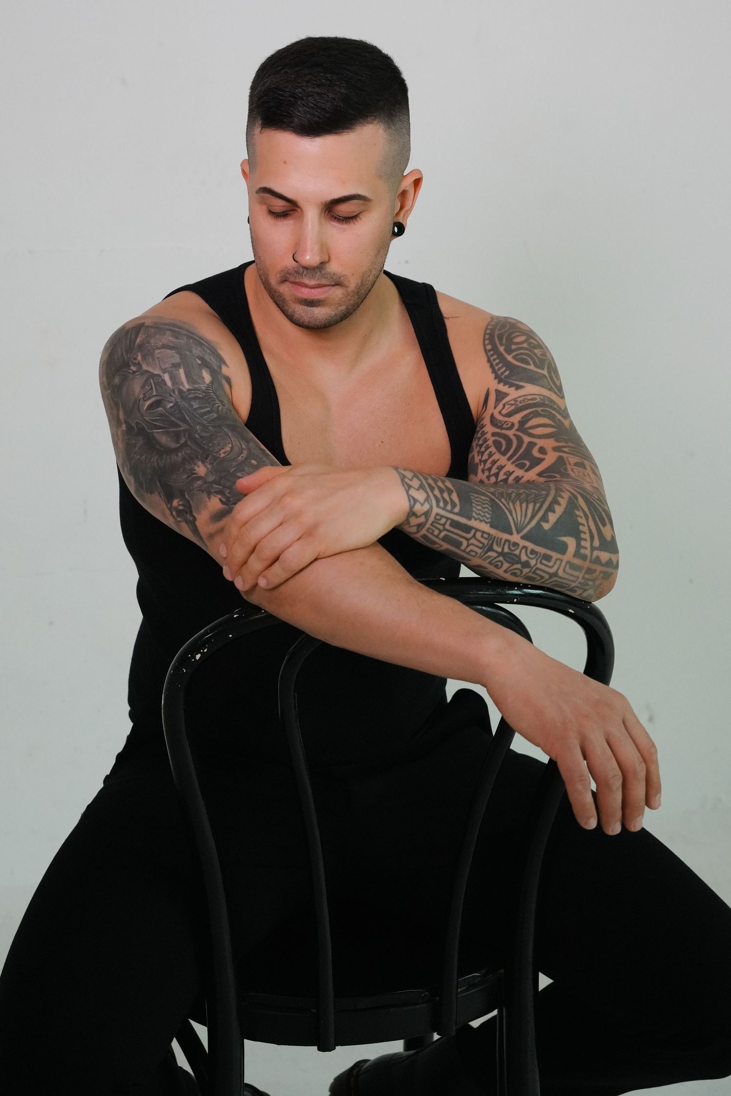 Muscular man with tattoos on both arms sits on black chair wearing sleeveless black outfit against white background.