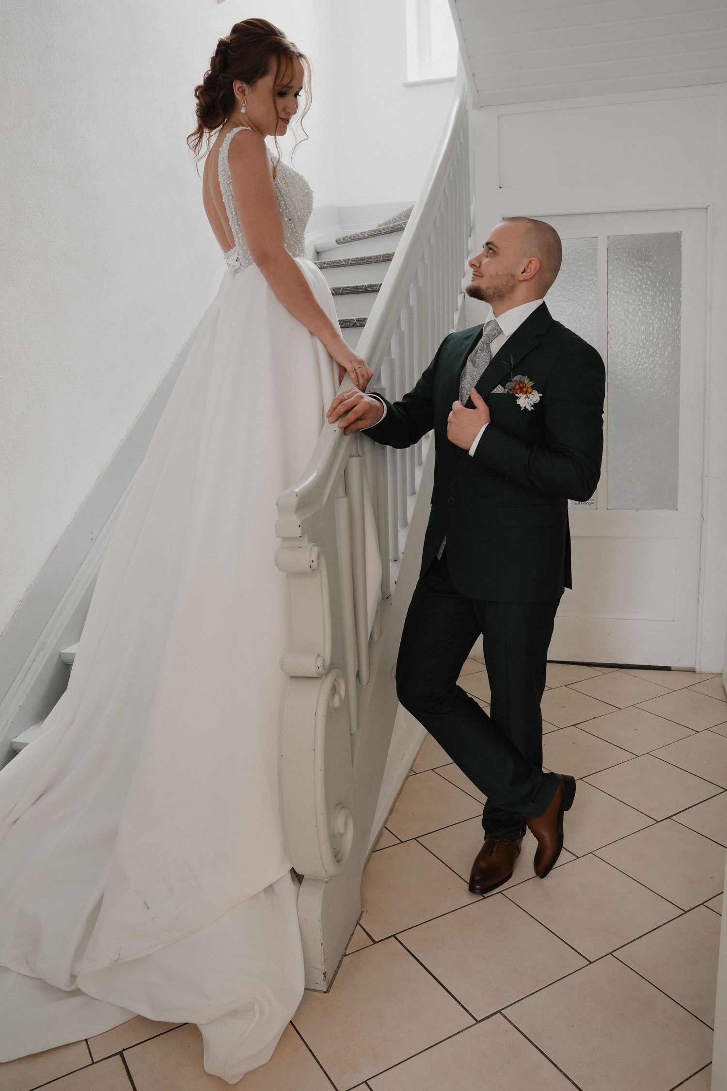 Groom in dark suit gazes up at bride in white gown on staircase, holding hands in romantic wedding portrait.