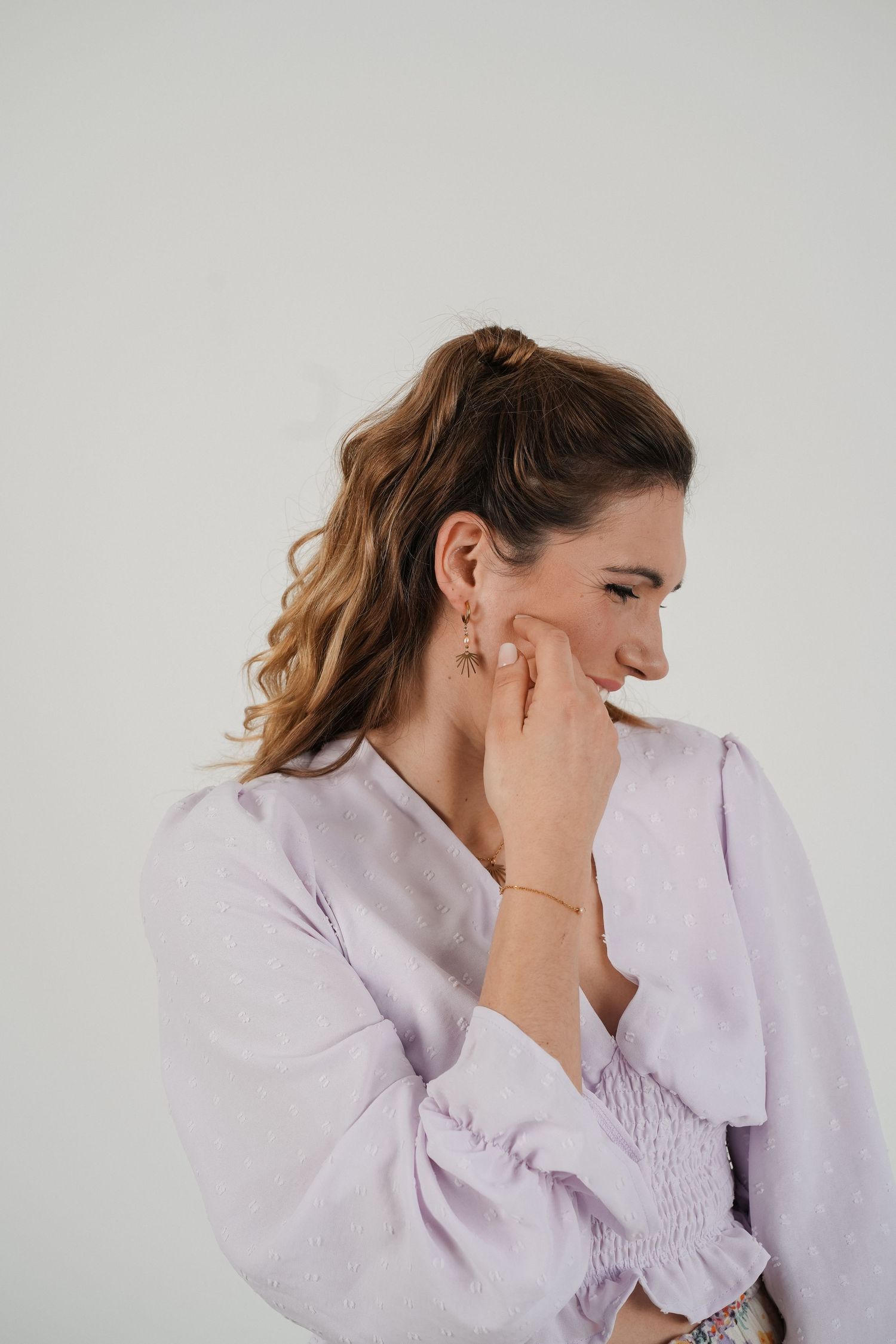 Woman in lavender blouse smiling, touching her gold hoop earring, with wavy ponytail on white background.