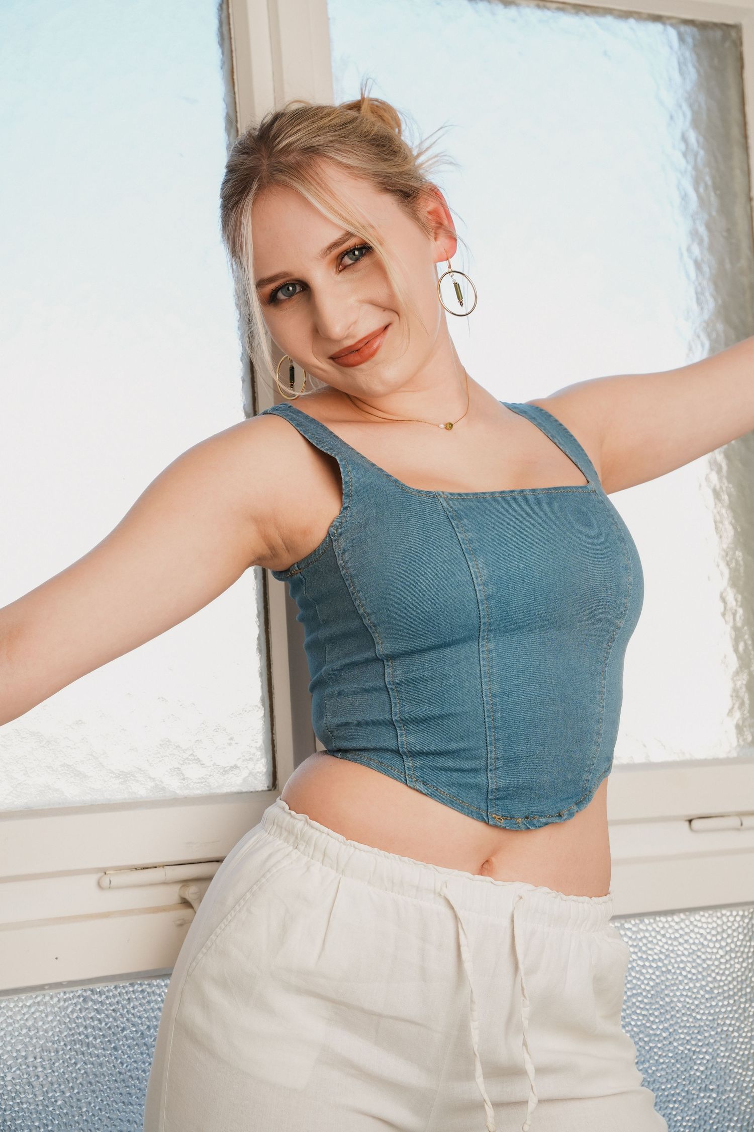 Blonde woman in denim crop top and white pants posing near a frosted window with hoop earrings.