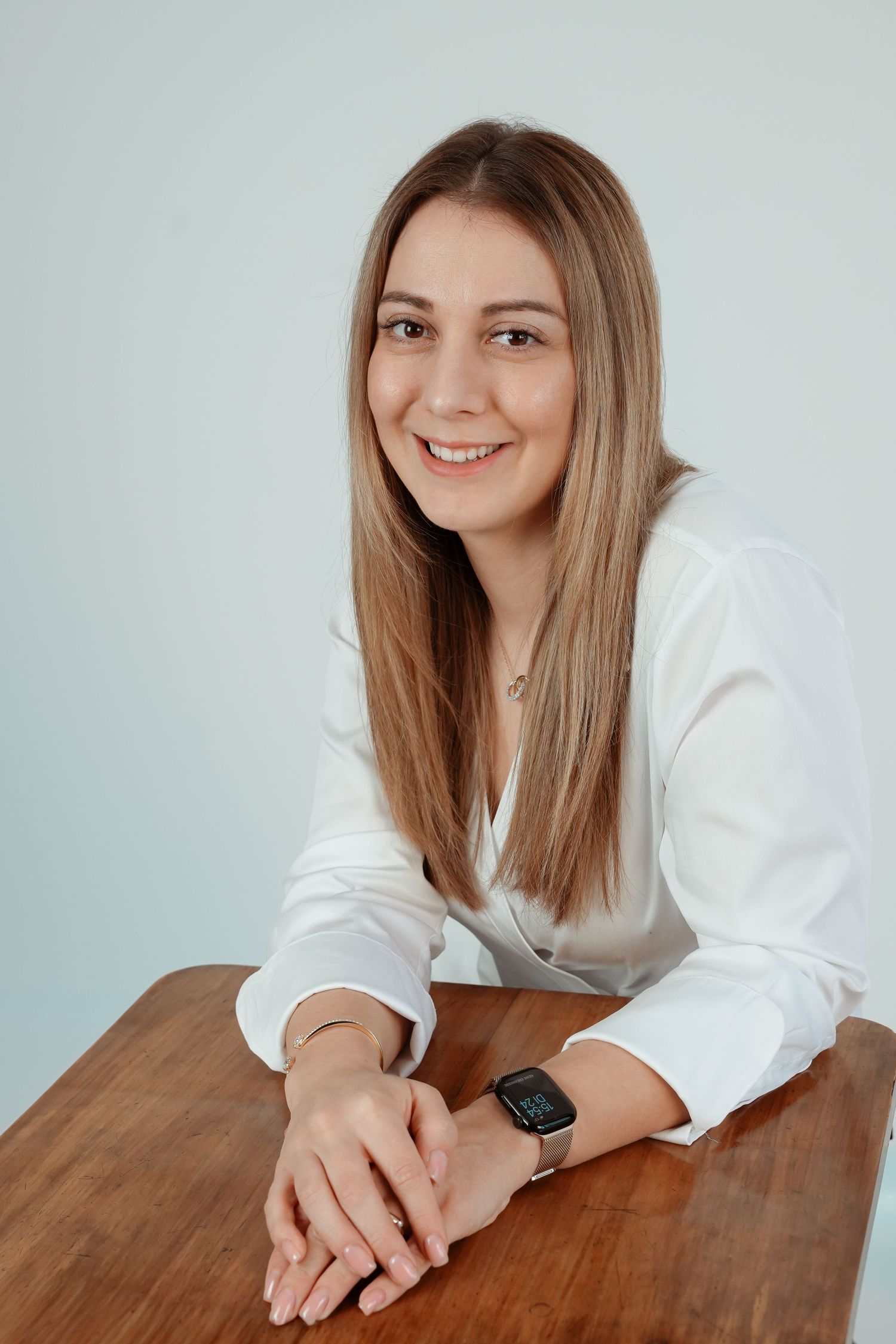 Young woman in white blouse smiling at desk, wearing smartwatch, with long blonde hair against light background.