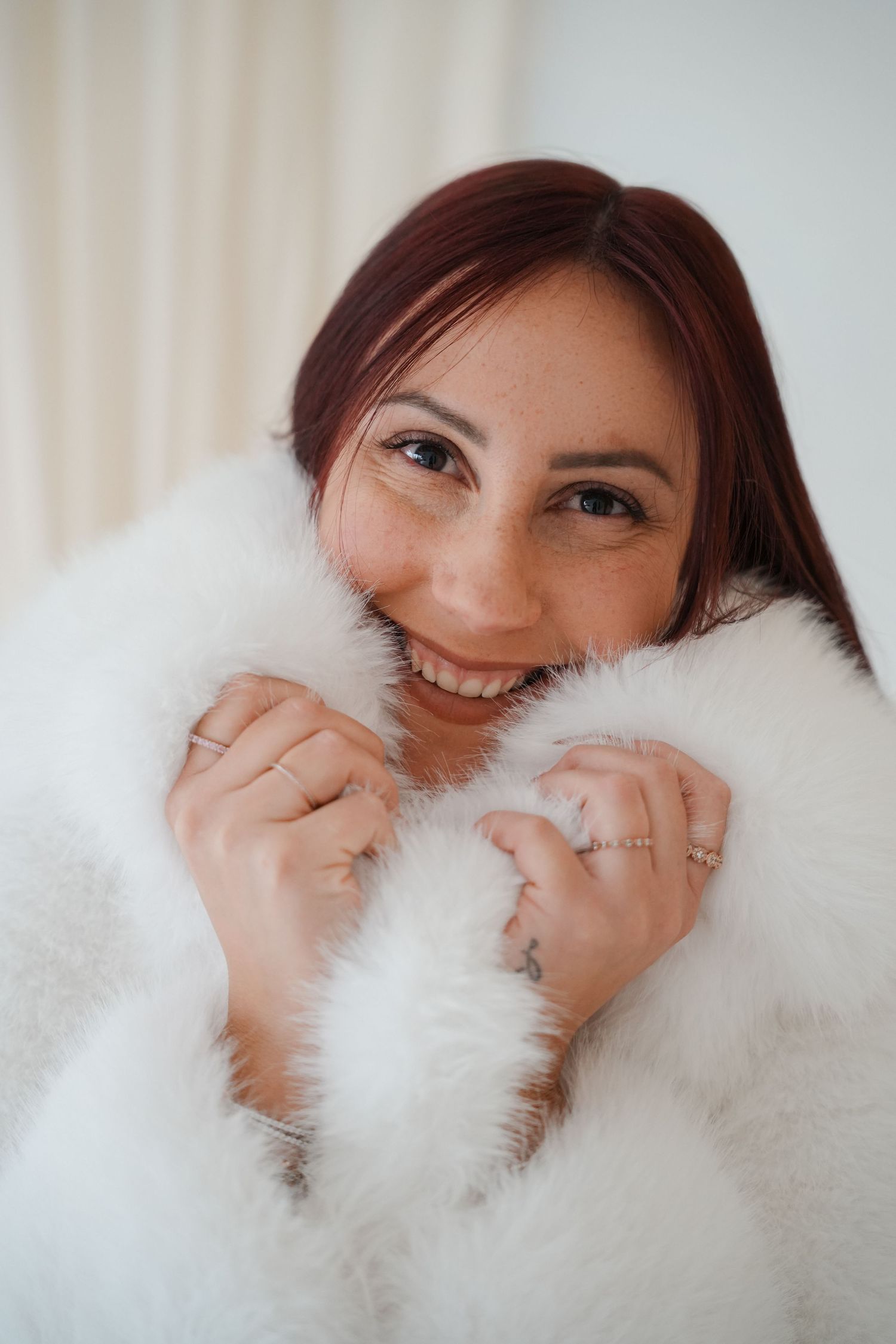 Smiling woman with red hair wrapped in a fluffy white fur coat, holding it close to her face.