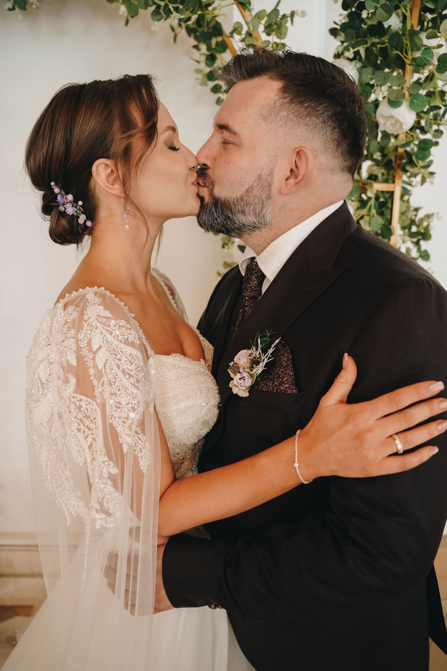 Bride and groom share a romantic first kiss at wedding ceremony, bride in lace gown, groom in dark suit with boutonniere.