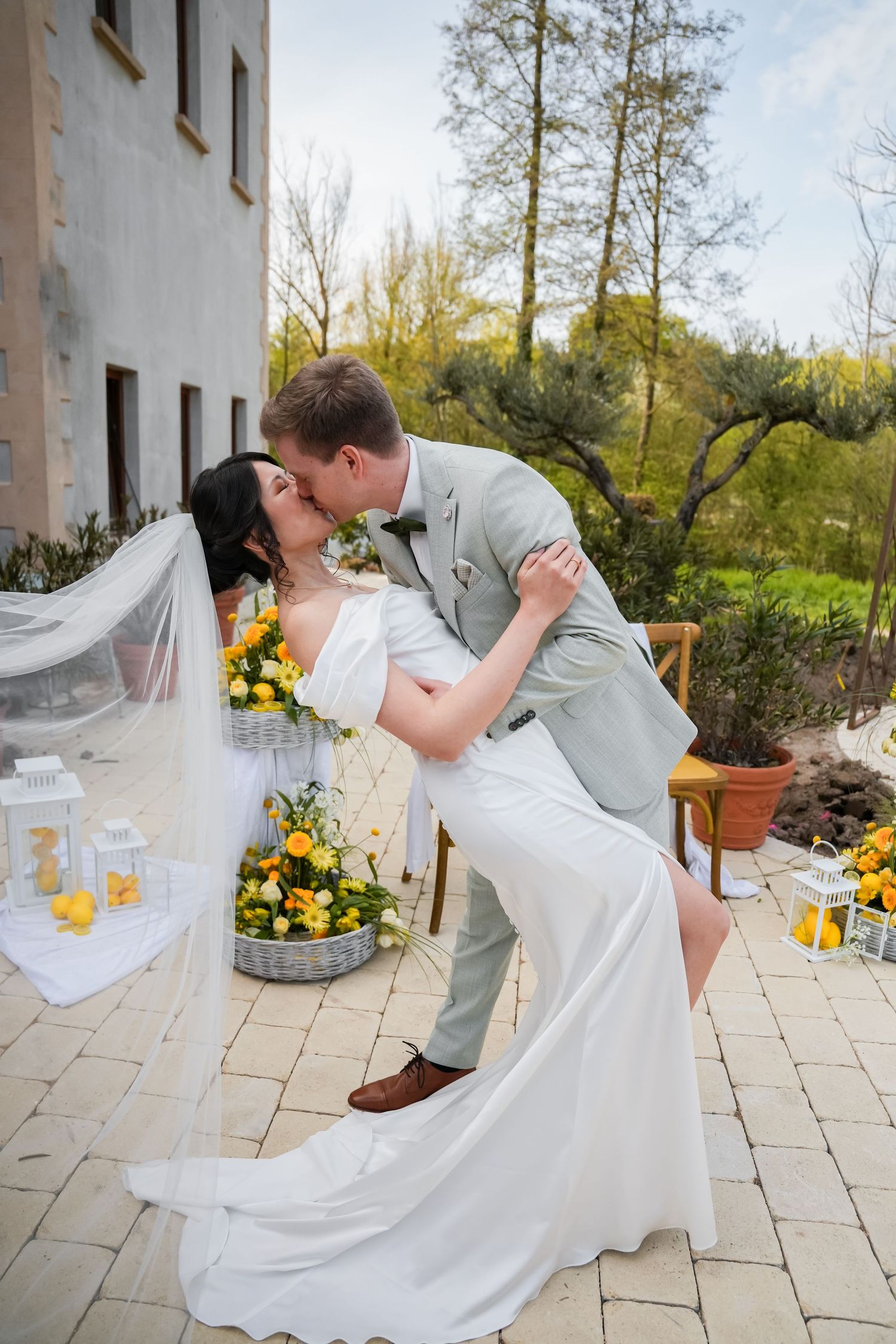 Bride and groom share a romantic dip kiss outdoors, surrounded by yellow floral arrangements and white lanterns.