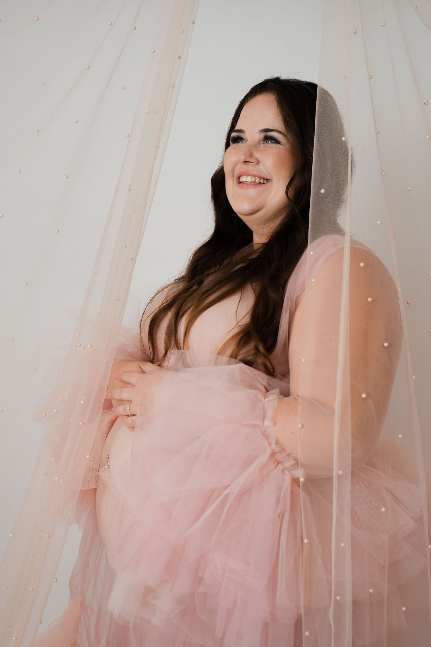Smiling pregnant woman in a pink tulle maternity gown holding her baby bump, posed against soft draped fabric backdrop.