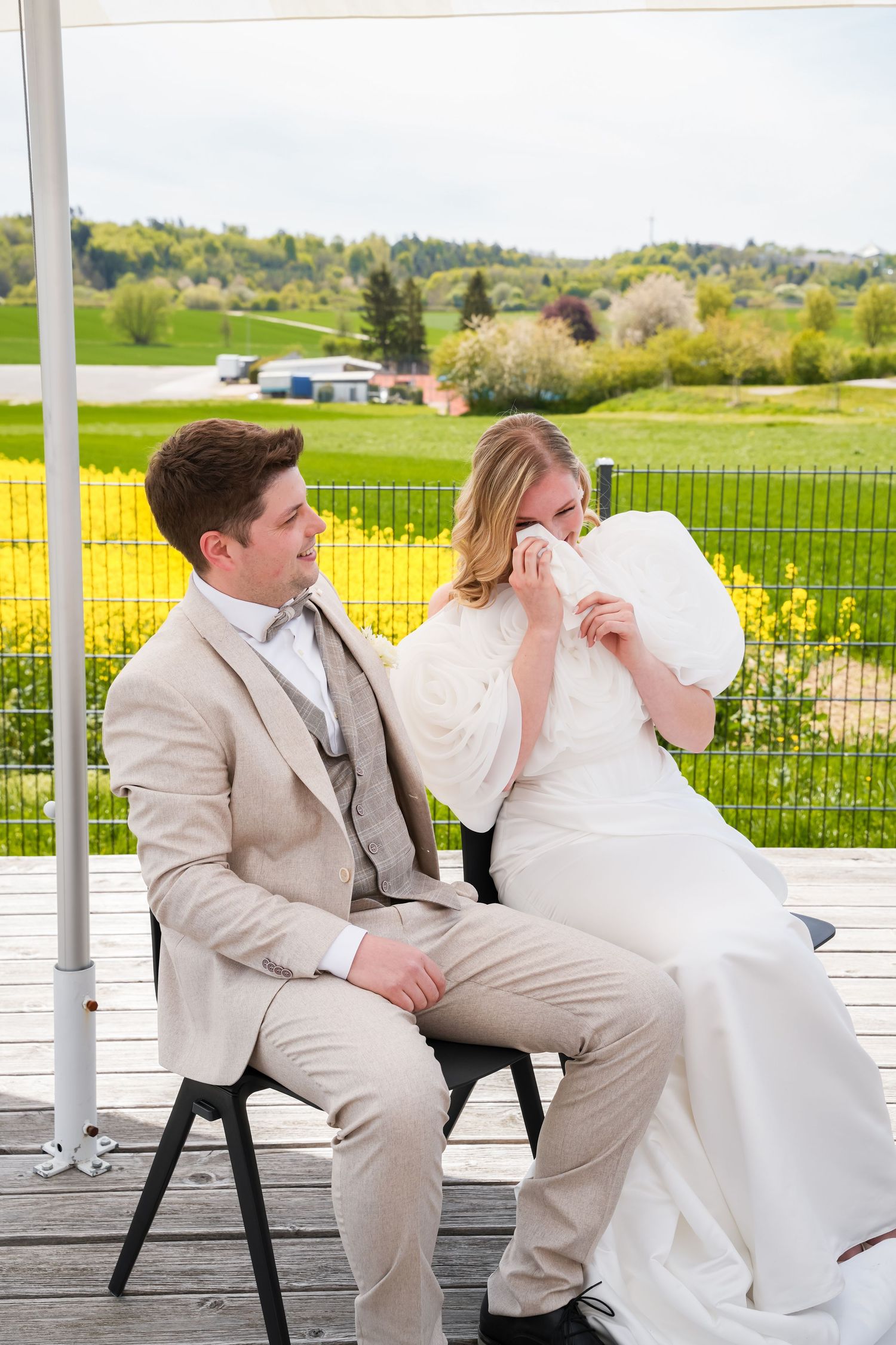 Bride wiping tears while groom sits beside her on a deck at an outdoor wedding ceremony with green landscape backdrop.