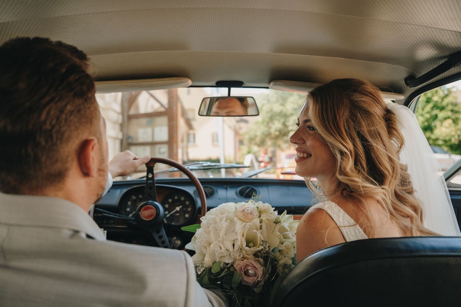 Bride holding bouquet smiles at groom while sitting in vintage car on wedding day.