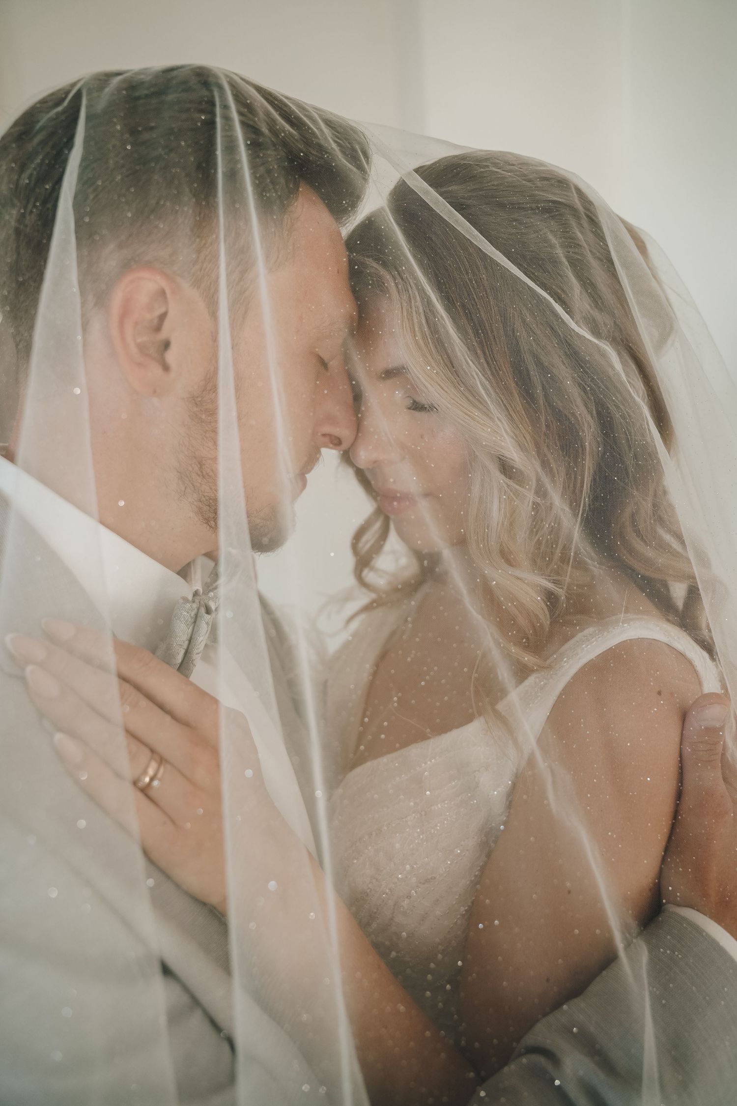 Bride and groom share an intimate moment under a wedding veil, foreheads touching, in a romantic soft-lit portrait.