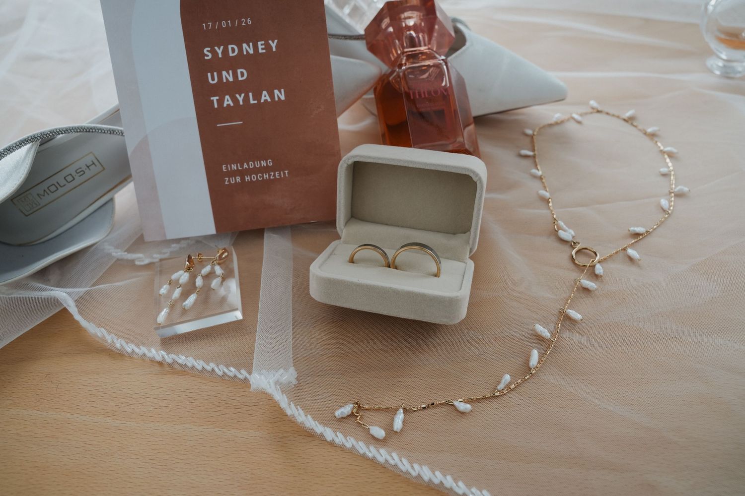 Wedding rings in a white box beside a floral necklace, earrings, and a Sydney und Taylan wedding invitation card.