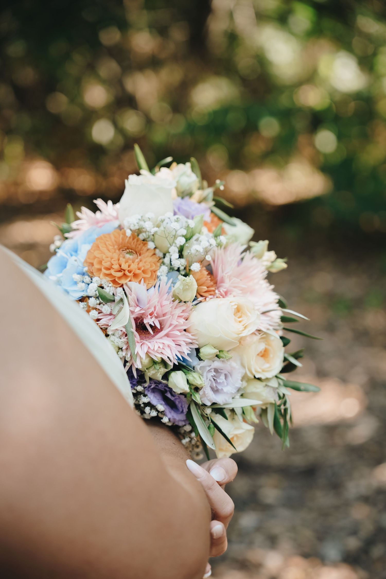 Bride holding a colorful wedding bouquet with pastel pink, orange, white, and lavender flowers outdoors.