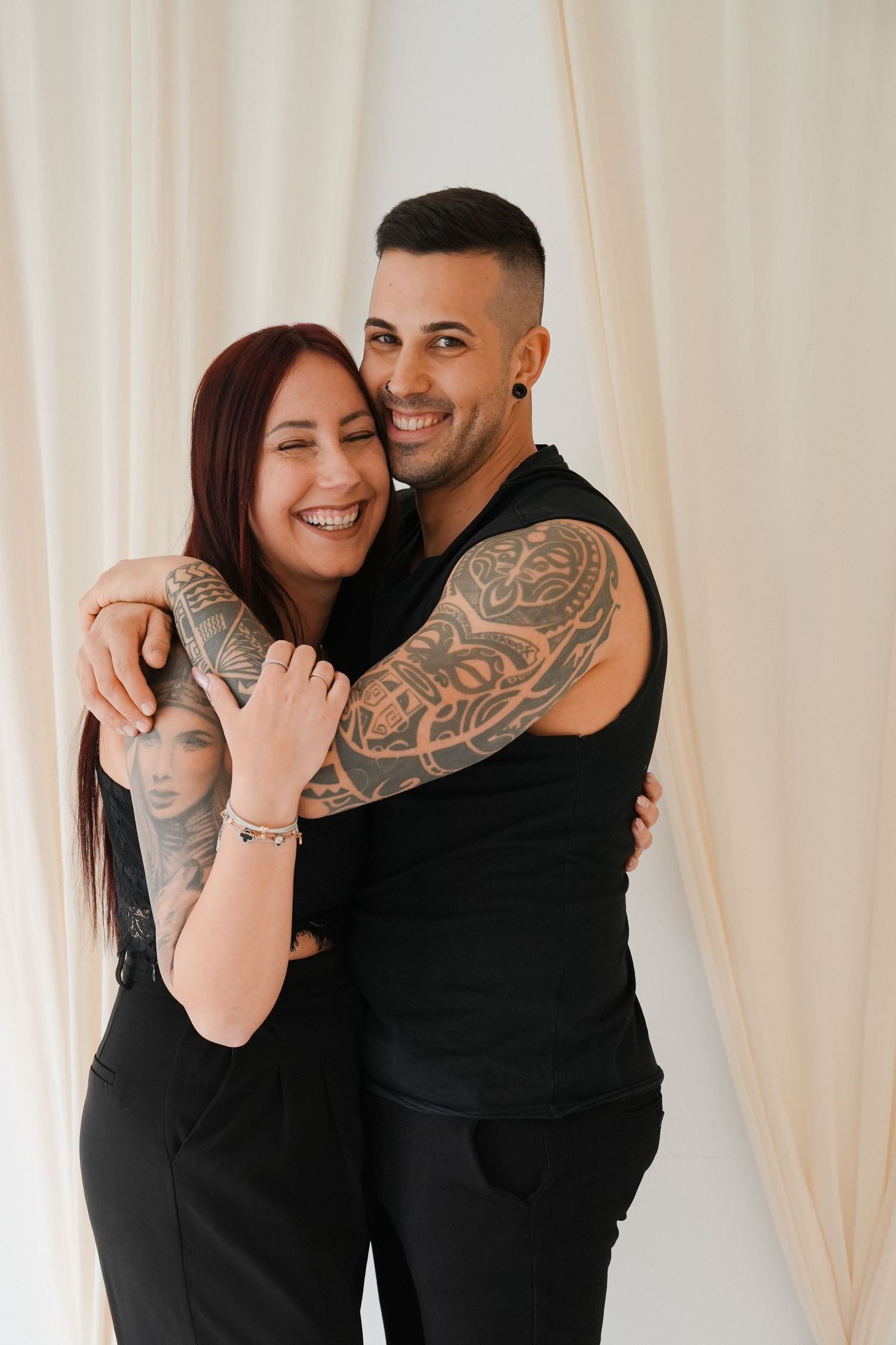 A smiling couple with tattoos wearing black outfits embrace warmly against a white curtain backdrop.