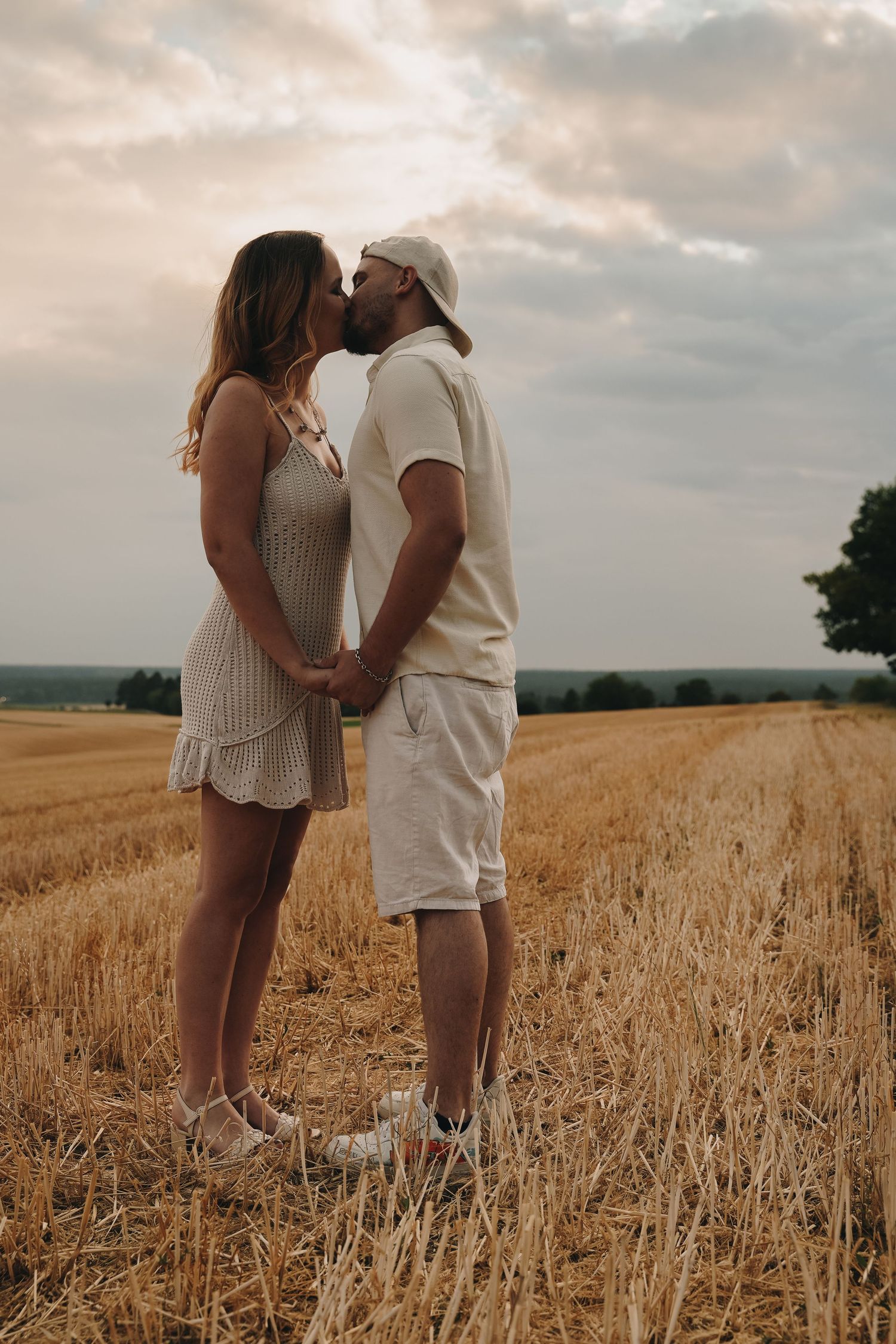 Couple kissing in a golden harvested field at sunset, holding hands, wearing casual summer outfits.