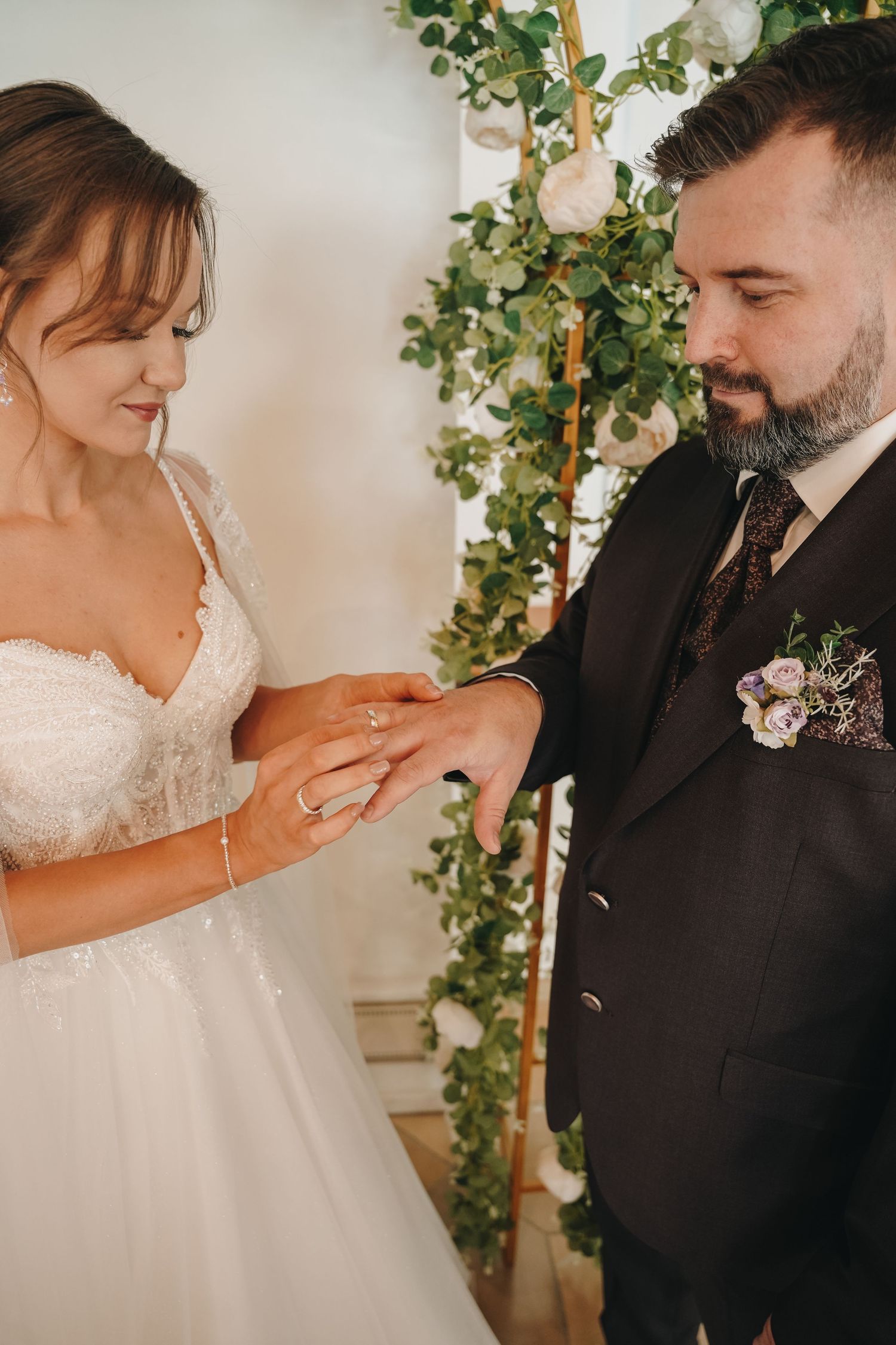 Bride placing a wedding ring on groom's finger during ceremony, with floral arch backdrop.