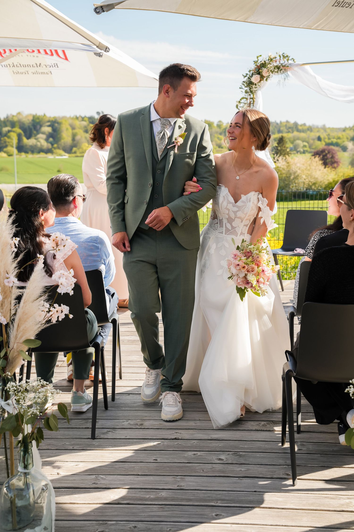 A joyful bride and groom walk down the aisle outdoors at a sunny wedding ceremony on a wooden deck.