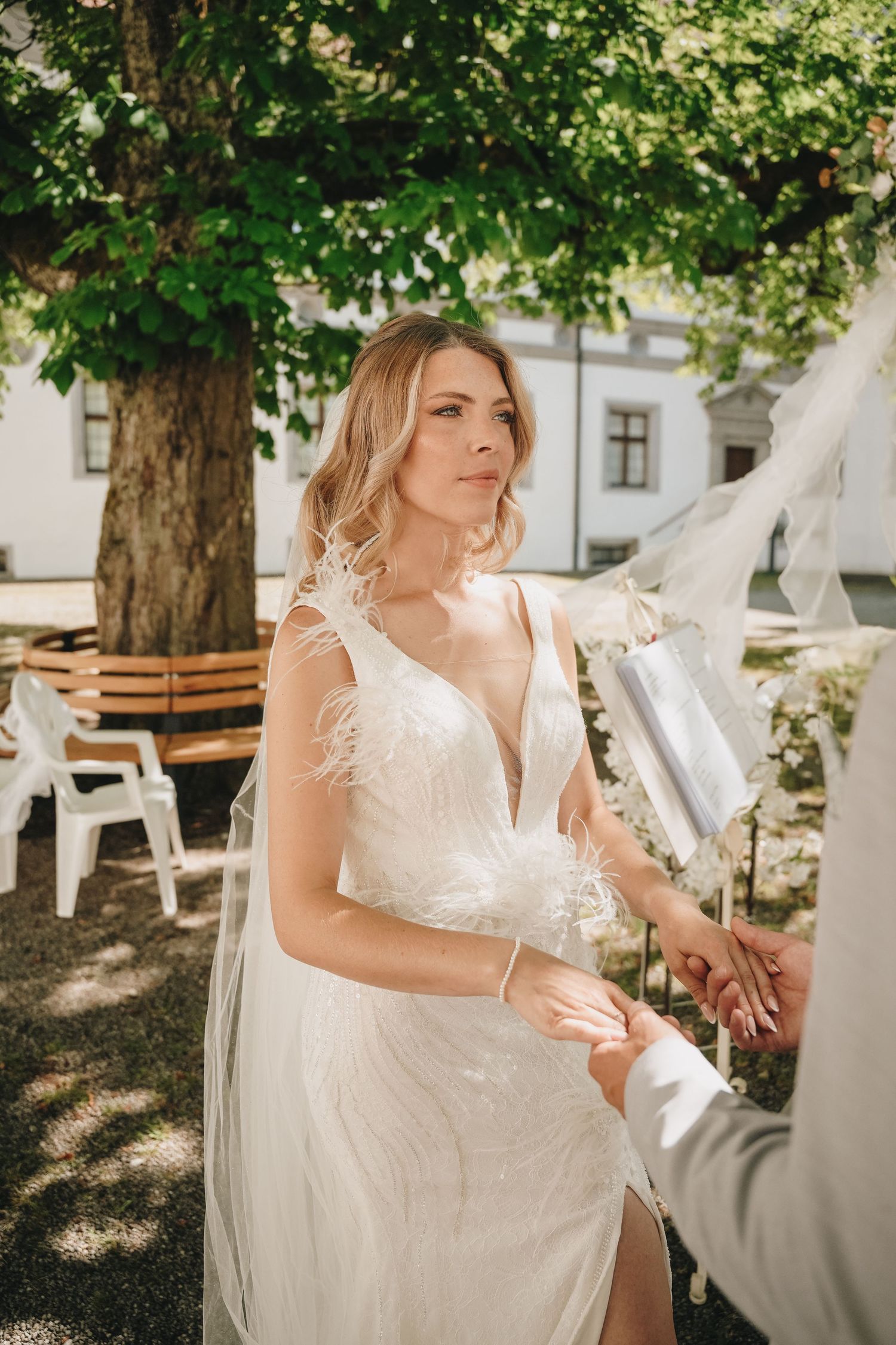 Elegant bride in a white feathered wedding gown holding hands outdoors under a green tree on a sunny day.