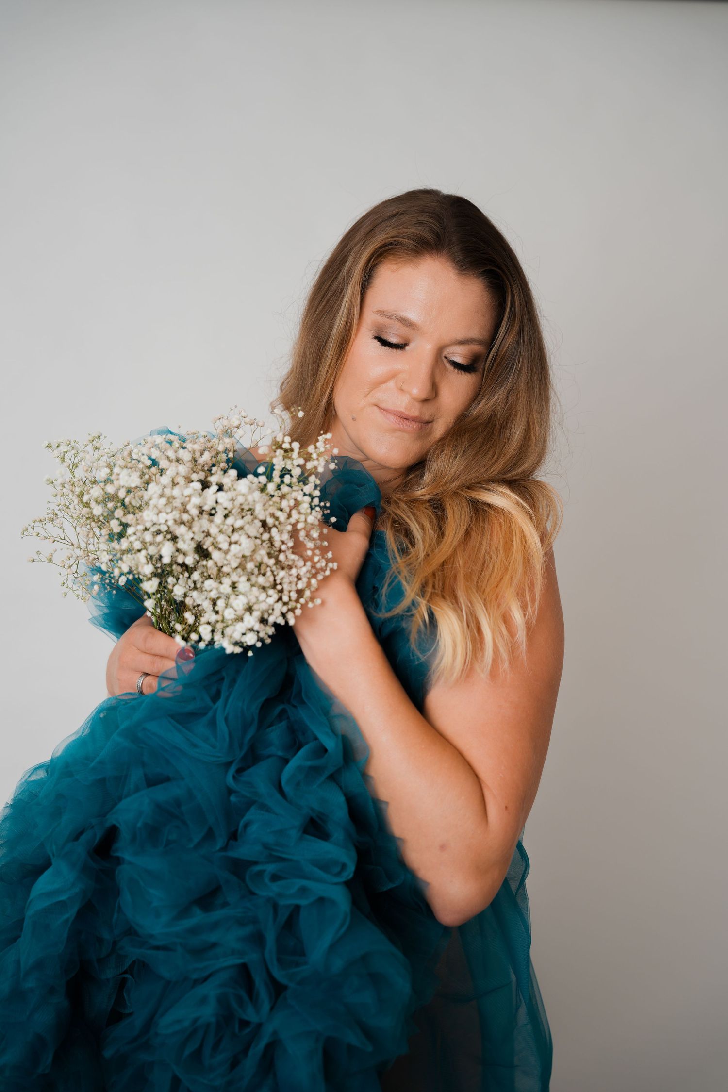 Woman in teal ruffled gown holding white baby's breath bouquet, eyes closed, against light gray background.