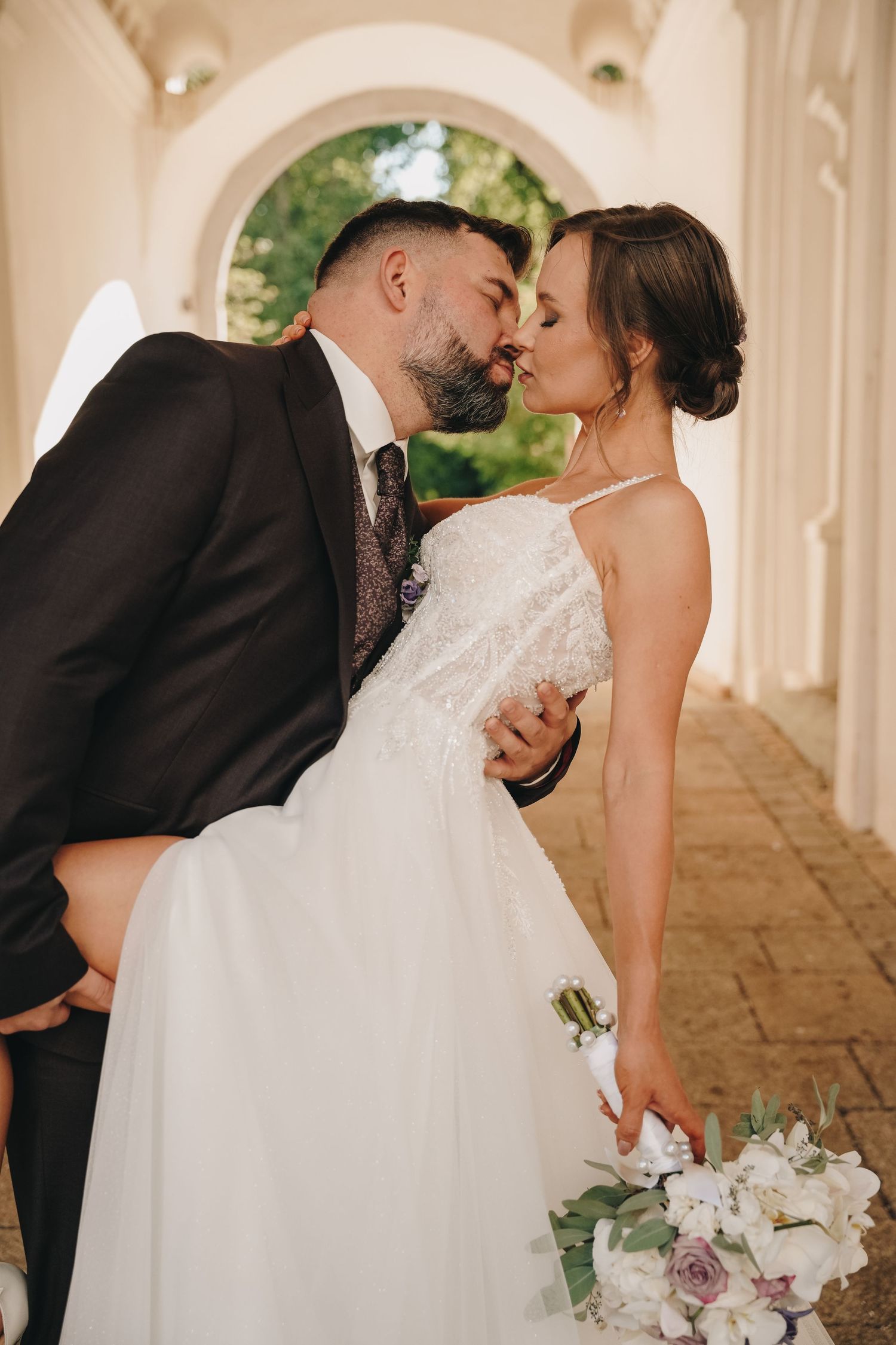 A bride in a lace wedding gown holding a white bouquet shares a kiss with her groom in a dark suit under a sunlit stone archway.