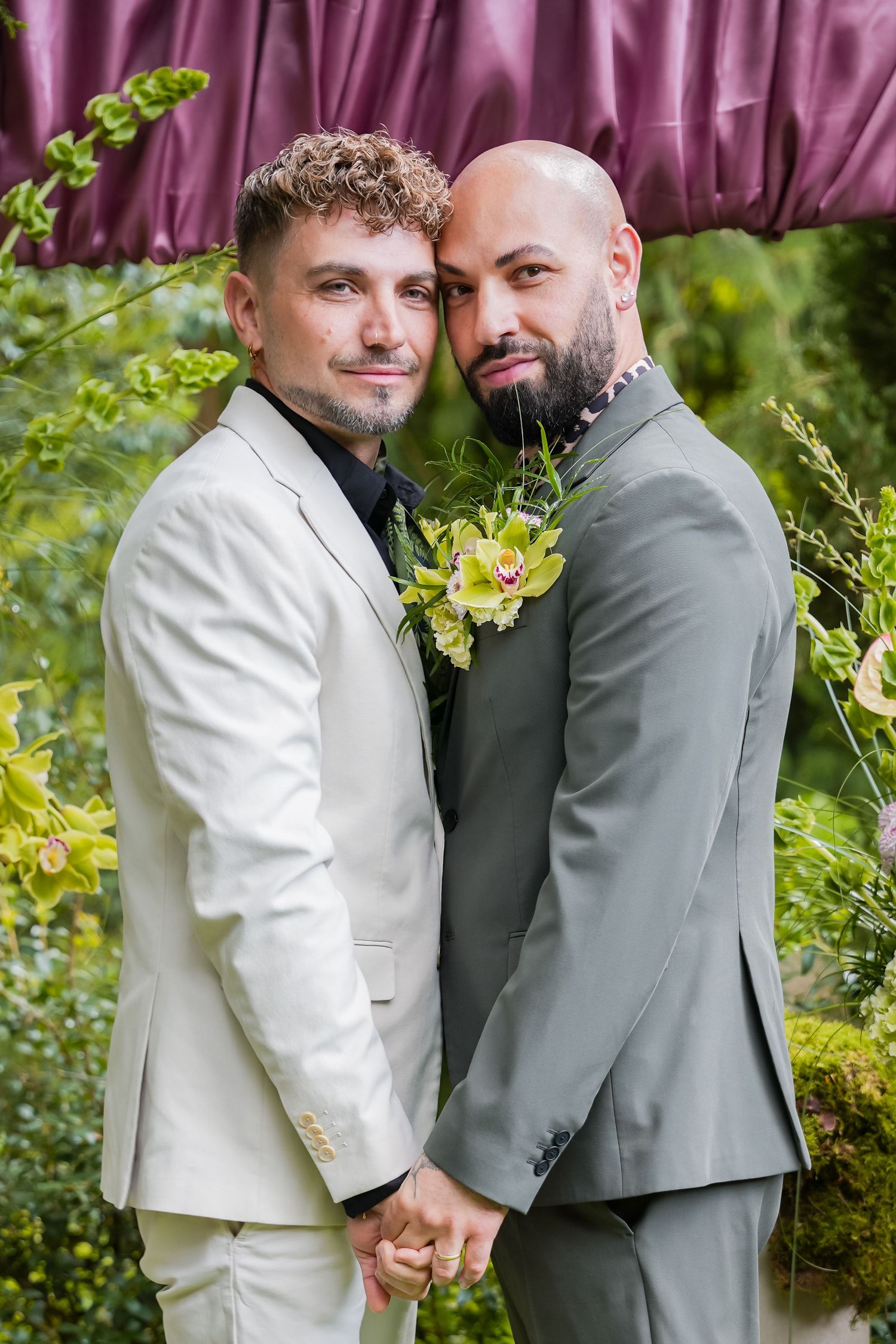 Two grooms in suits holding hands at their wedding ceremony, surrounded by lush greenery and a purple canopy.