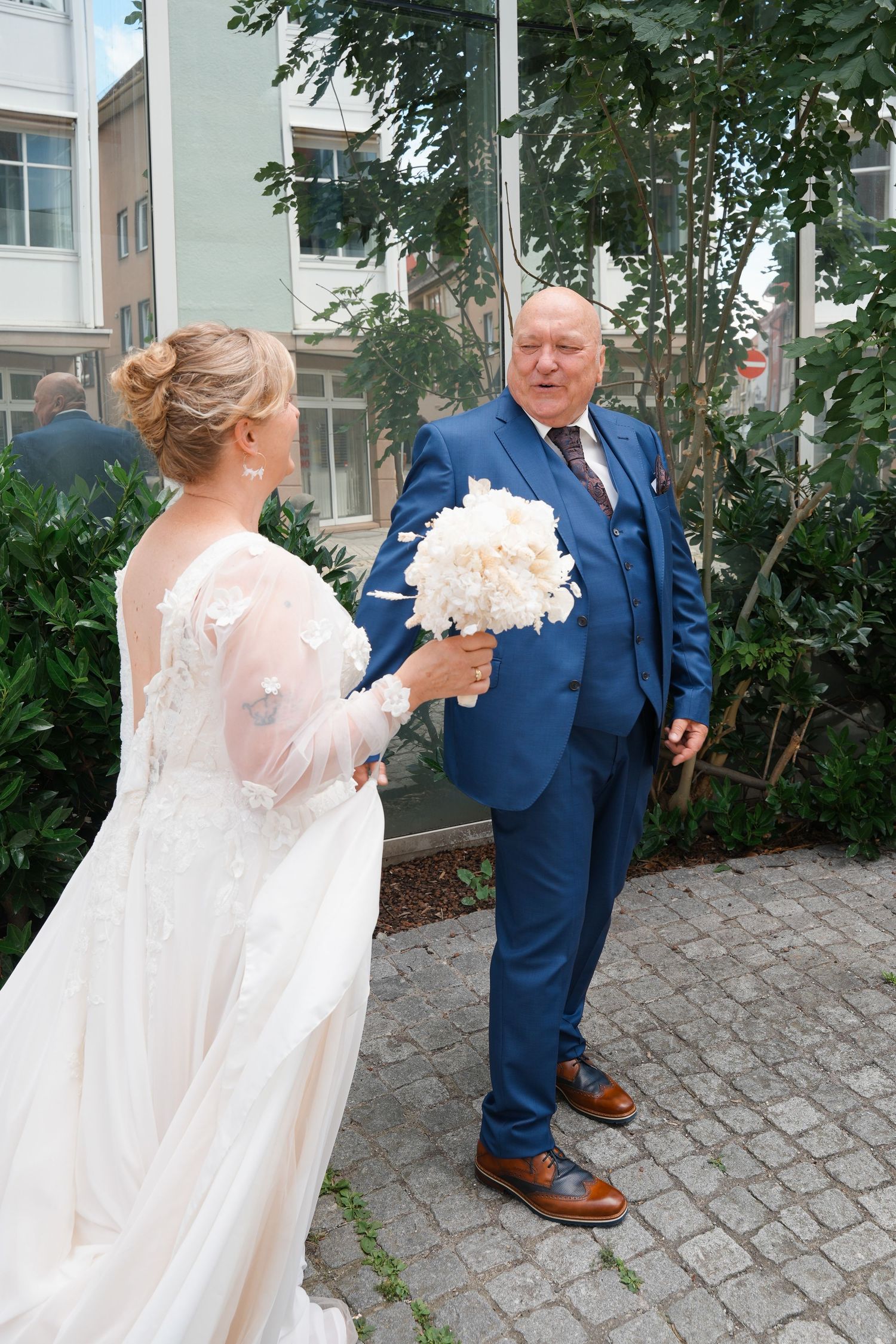 Bride in white lace gown holds bouquet while groom in blue suit smiles outdoors at wedding ceremony.