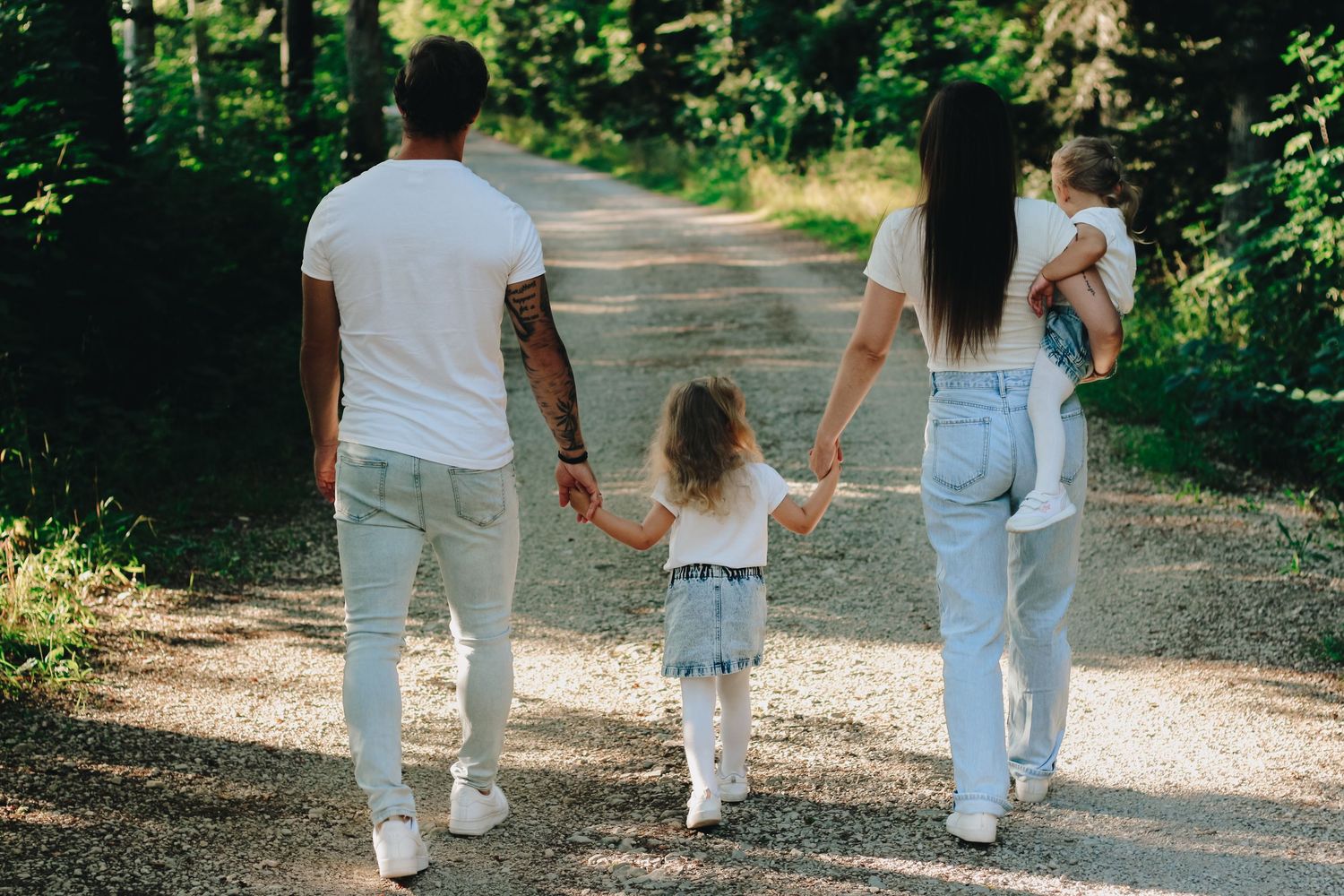Family of four walking hand-in-hand down a scenic forest path, parents with two young children in casual white outfits.
