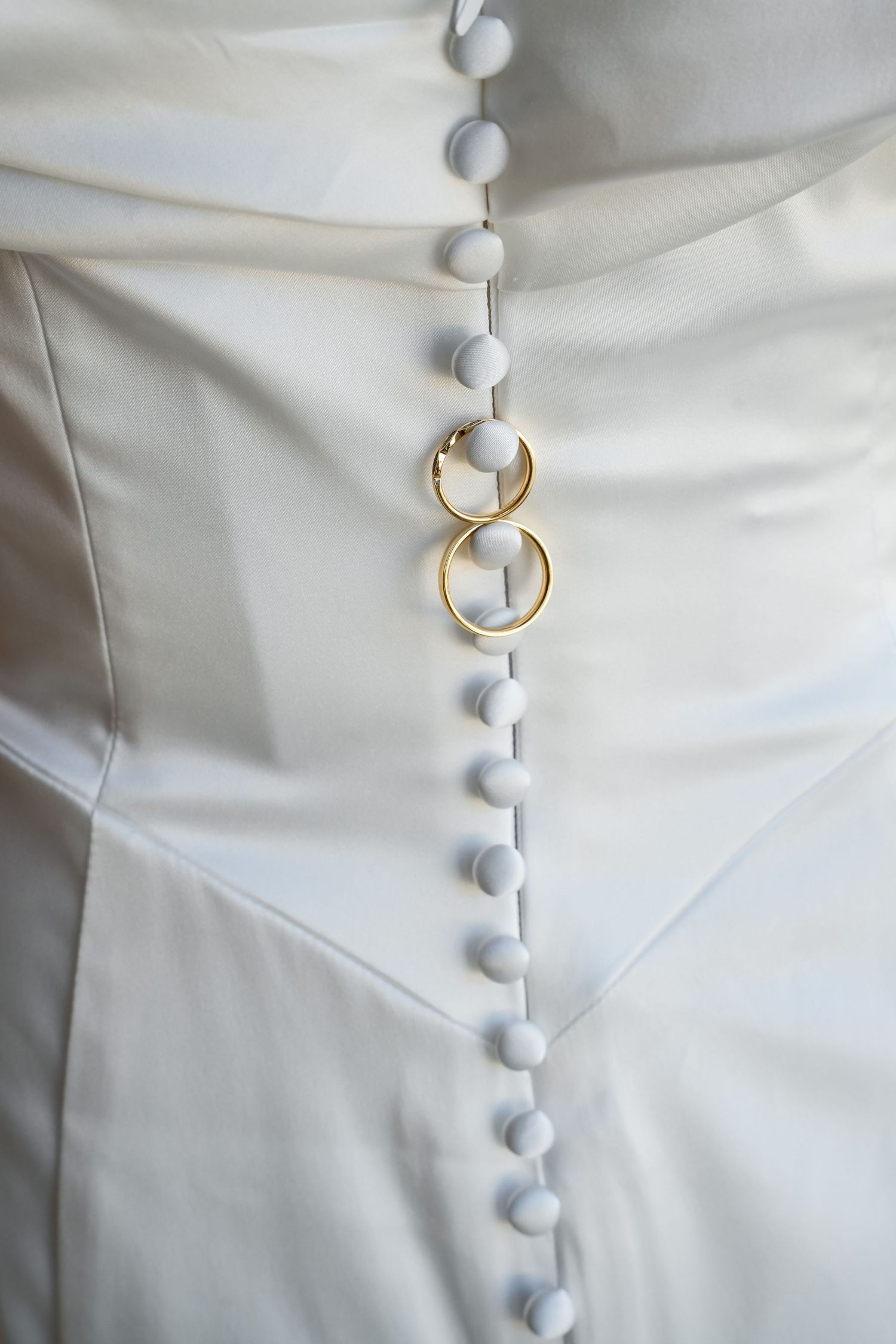 Two gold wedding rings resting on white satin bridal gown buttons, symbolizing marriage and elegance.