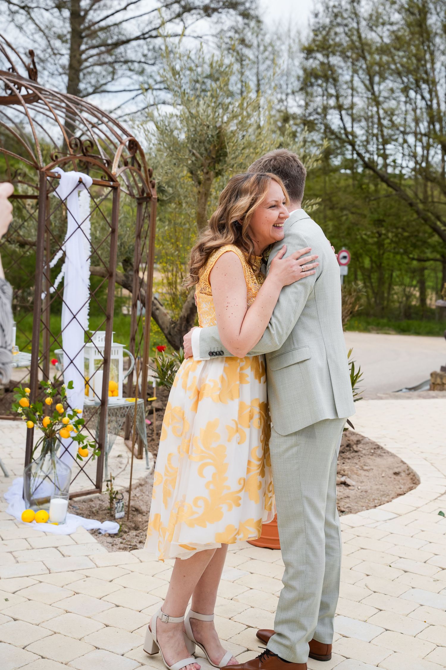 A woman in a yellow floral dress hugs a man in a light gray suit at an outdoor wedding ceremony.