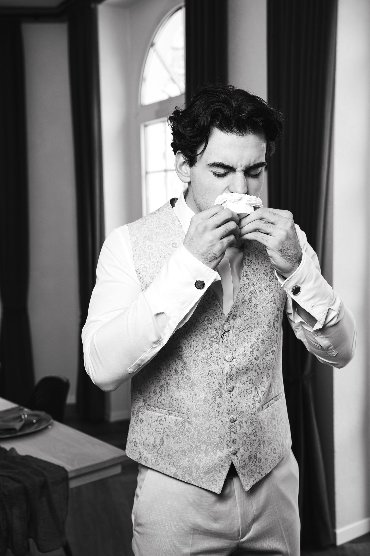 Groom in patterned vest smelling boutonniere in black and white wedding preparation photo.