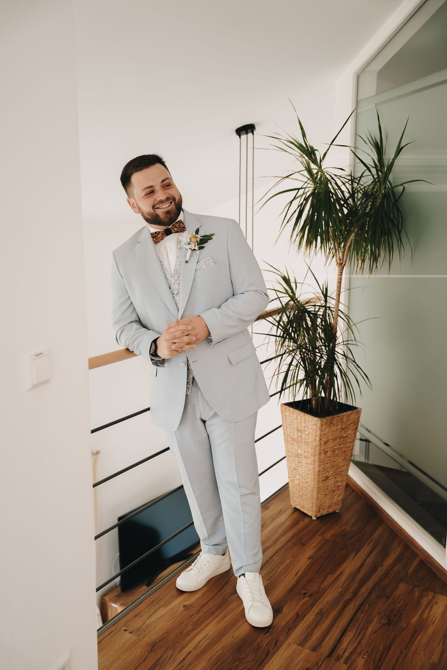 Smiling groom in light gray suit with boutonniere and bow tie posing indoors beside a tall green plant.