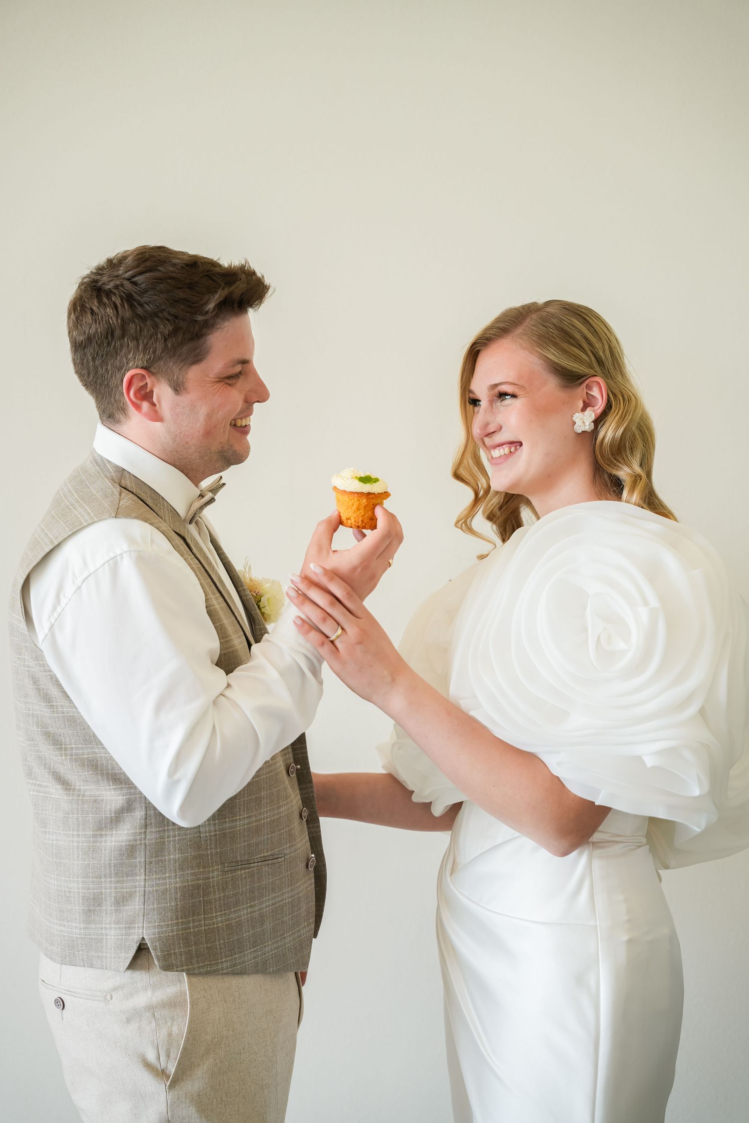 Bride and groom smiling while sharing a cupcake on their wedding day, dressed in elegant white and grey attire.