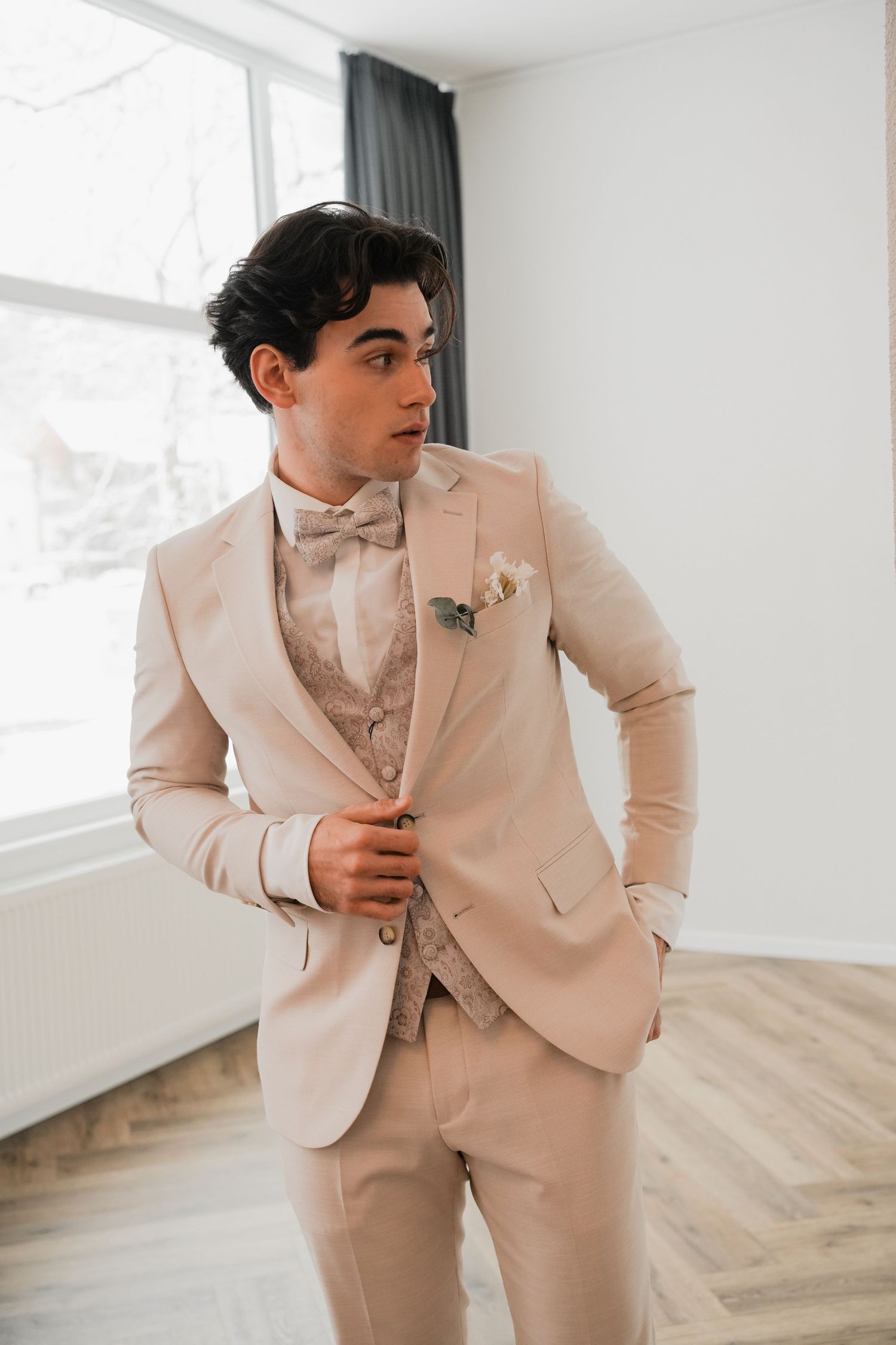 Young man in beige three-piece suit with bow tie and boutonniere, posing elegantly in bright room.