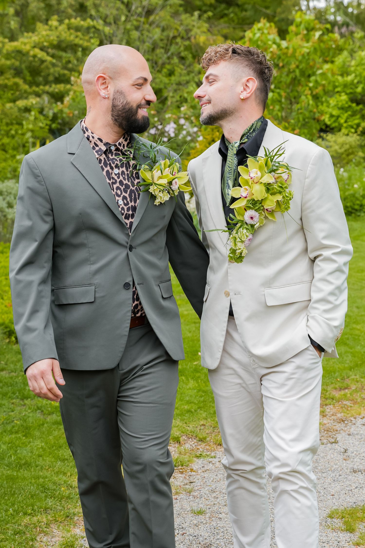 Two grooms in matching gray suits with floral boutonnieres smile at each other outdoors at their wedding.