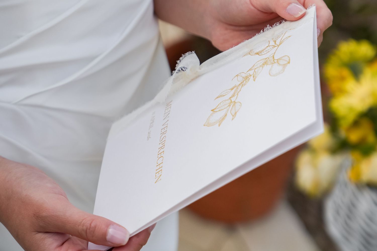 Hands holding an elegant white wedding card with gold floral design, with yellow flowers in the background.