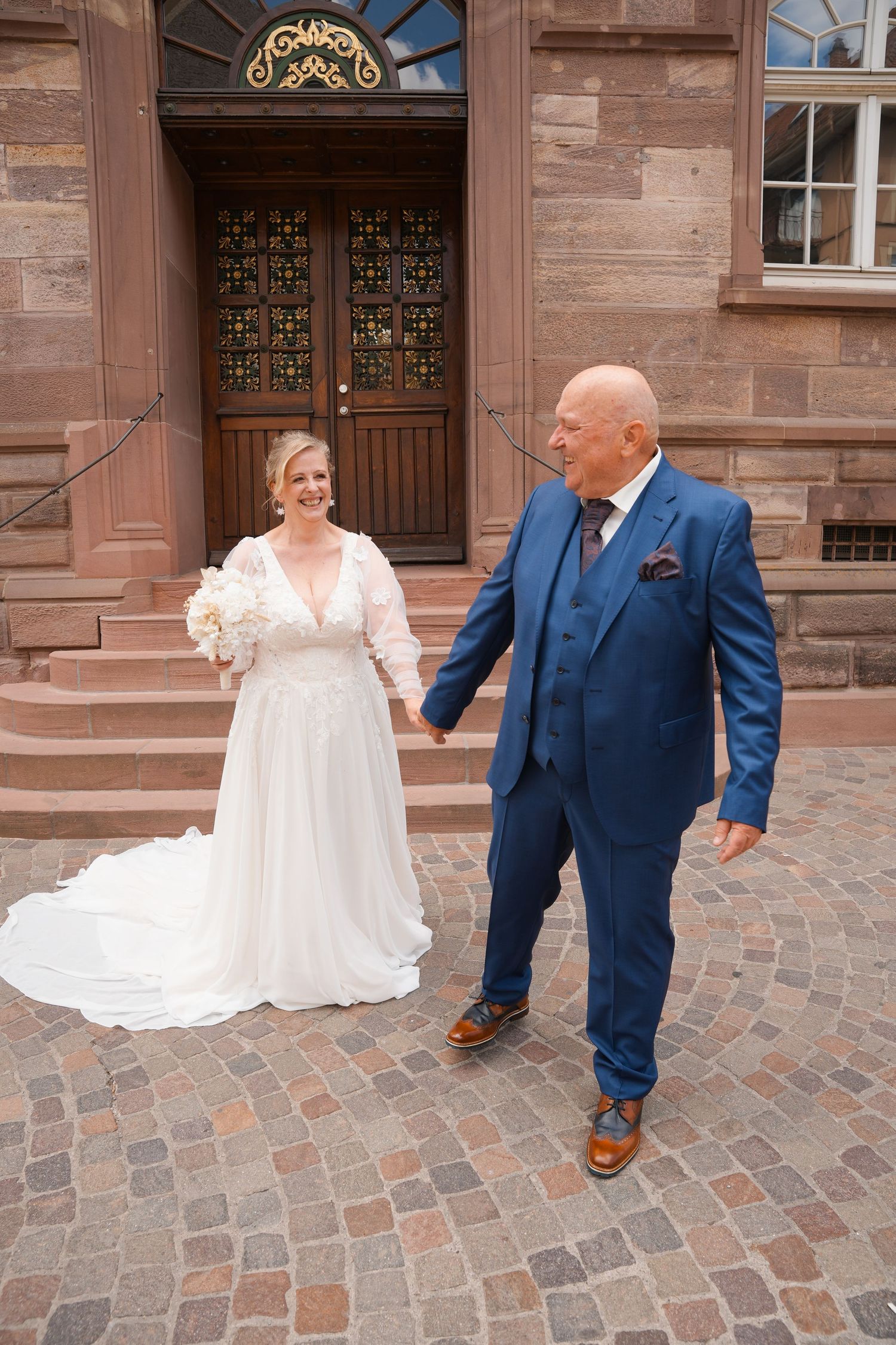 Bride in white gown and groom in blue suit hold hands on cobblestone plaza outside historic building.