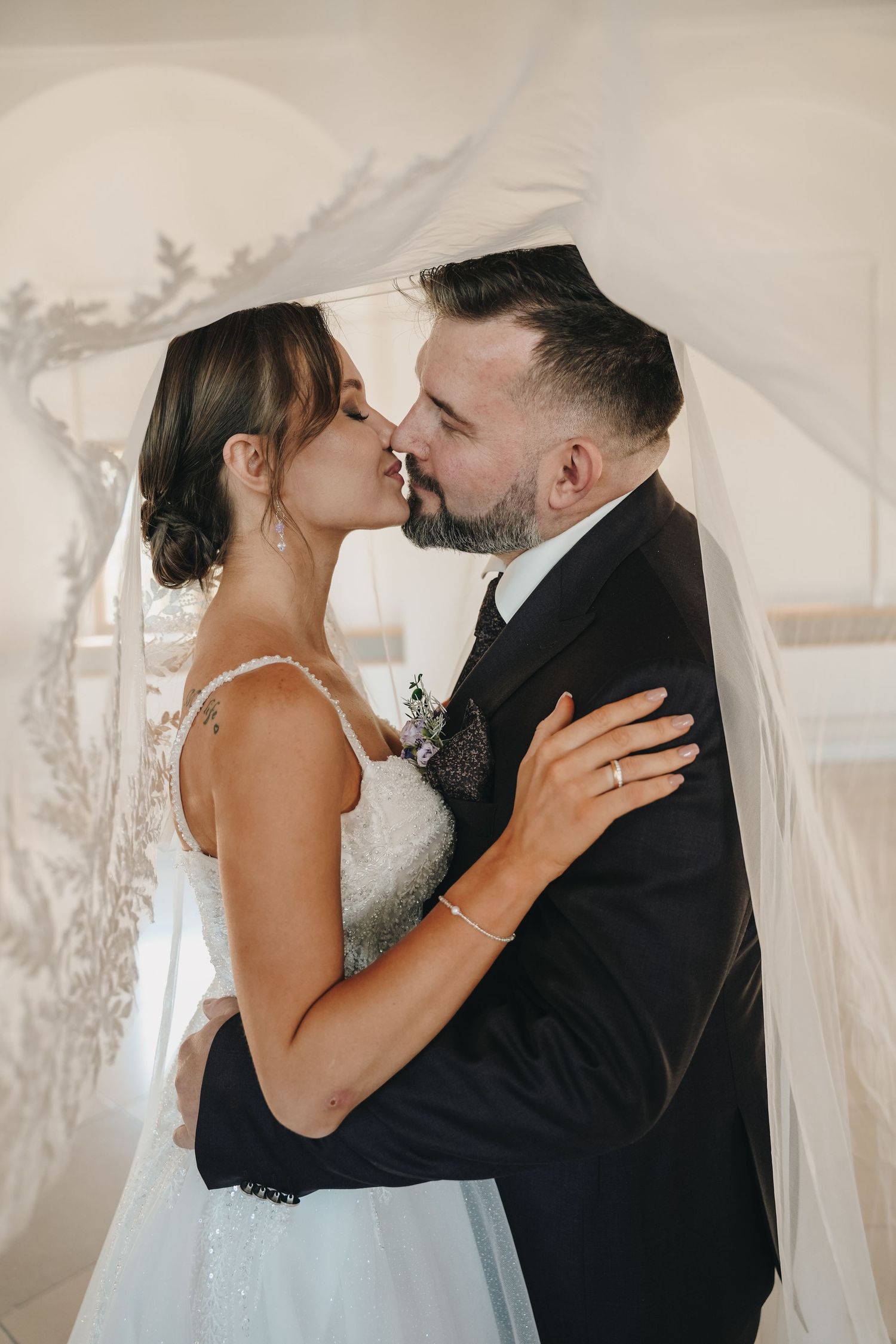 Bride and groom share a romantic kiss under a wedding veil, dressed in elegant bridal gown and dark suit.