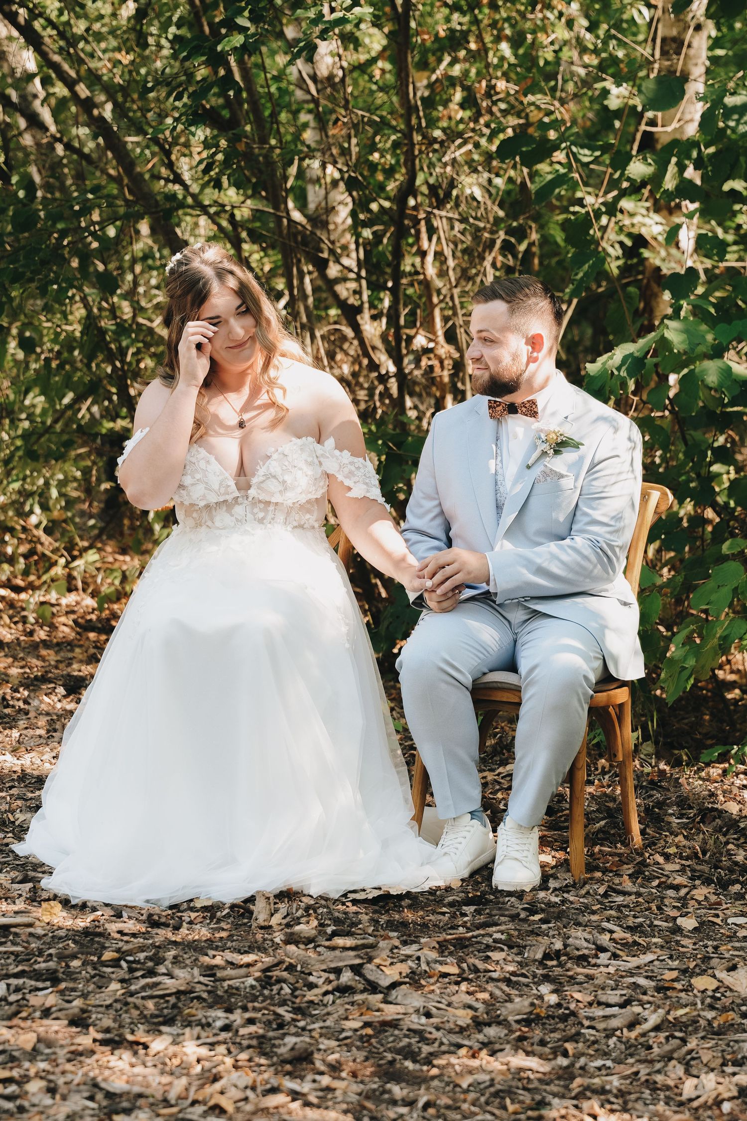 Bride in white off-shoulder gown and groom in light blue suit hold hands on chairs outdoors.