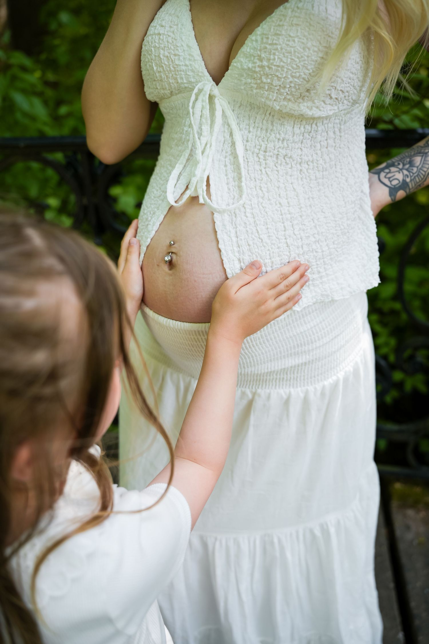 Young girl touching pregnant woman's bare belly bump outdoors, both wearing white dresses in garden setting.