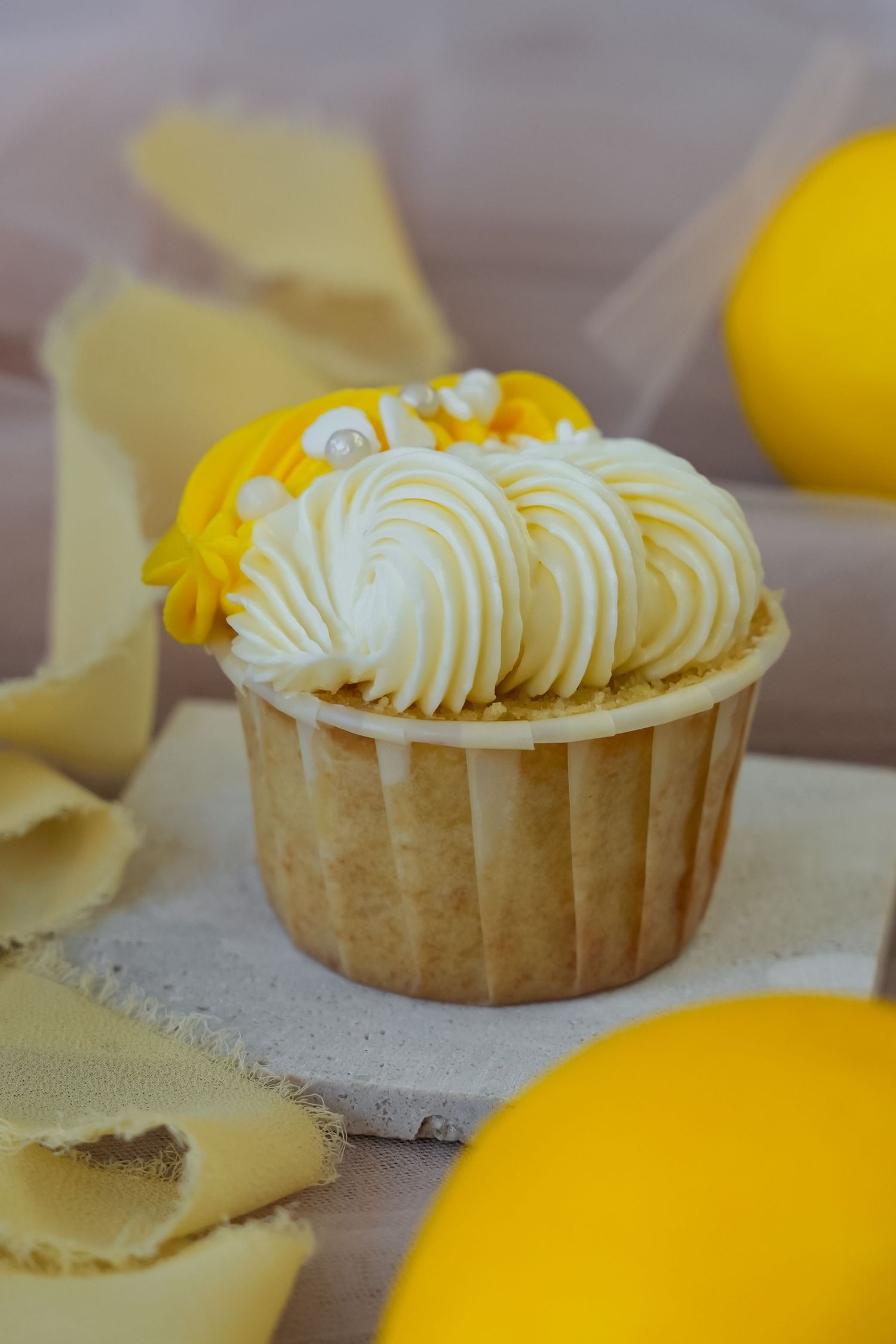 Lemon cupcake with swirled white frosting, yellow flower decoration, and pearl sprinkles in a gold wrapper.