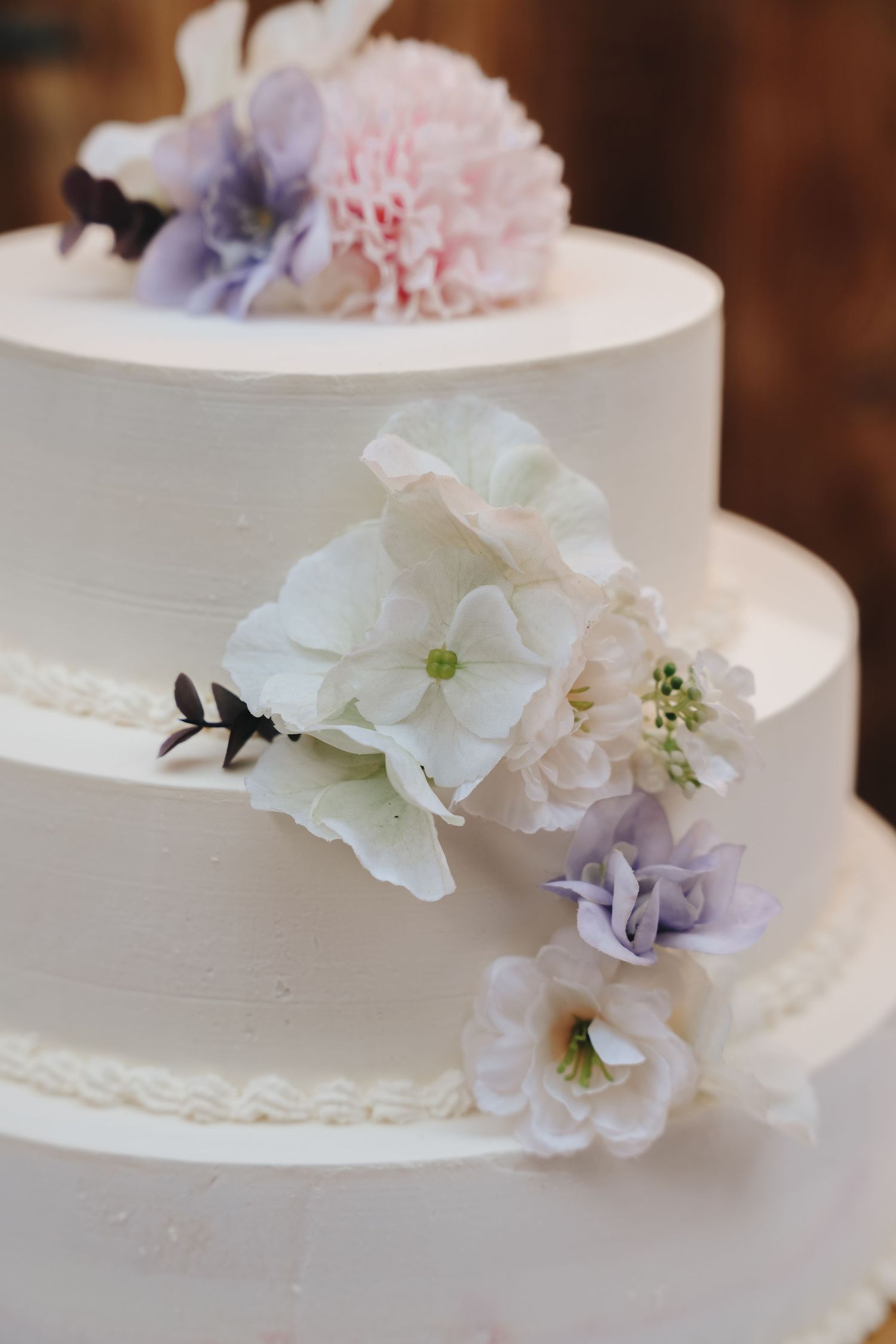 Elegant three-tier white wedding cake decorated with pastel pink, lavender, and white flowers.