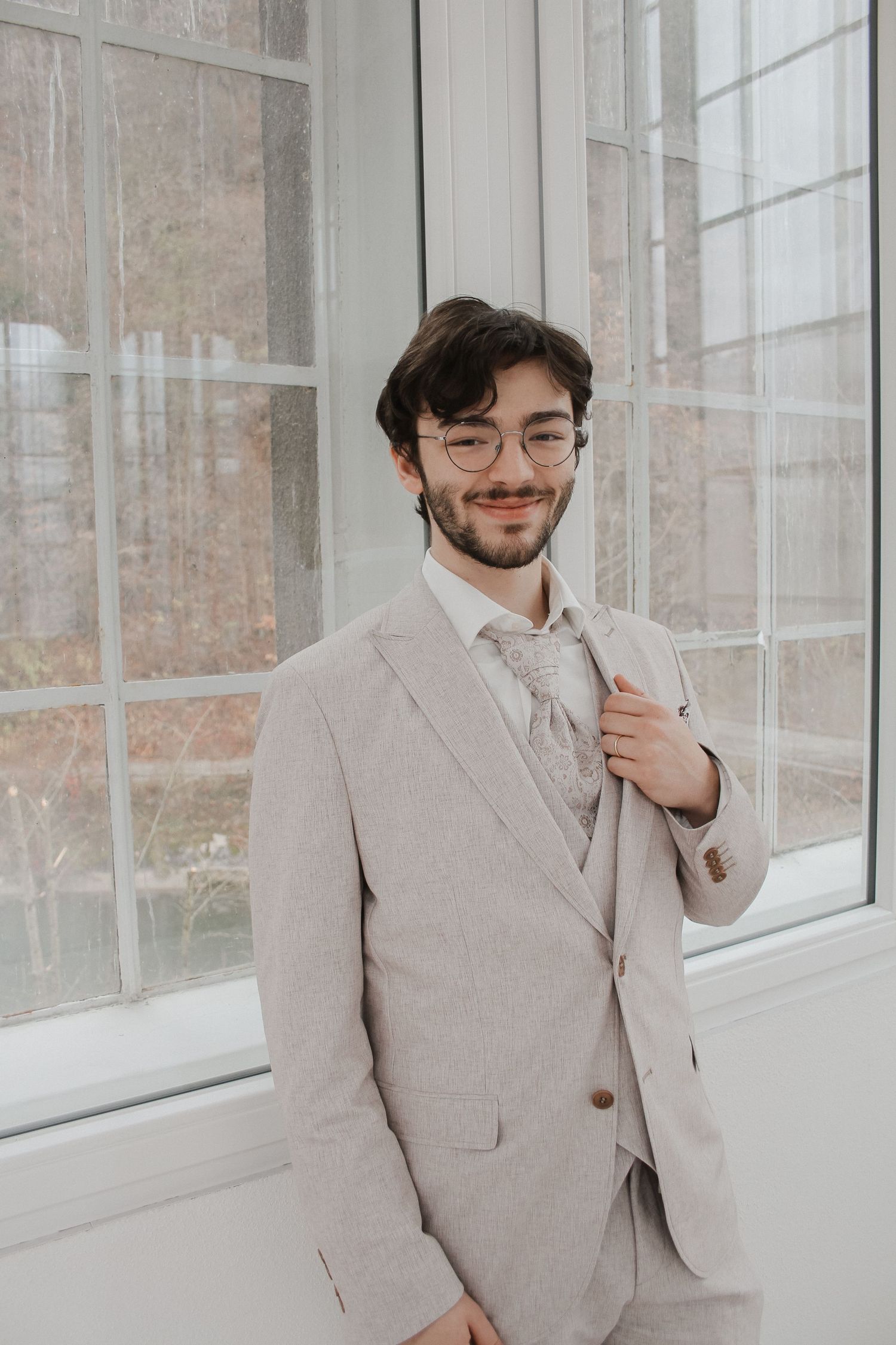 Young man in beige suit and glasses smiling near bright windows, dressed for a formal occasion.