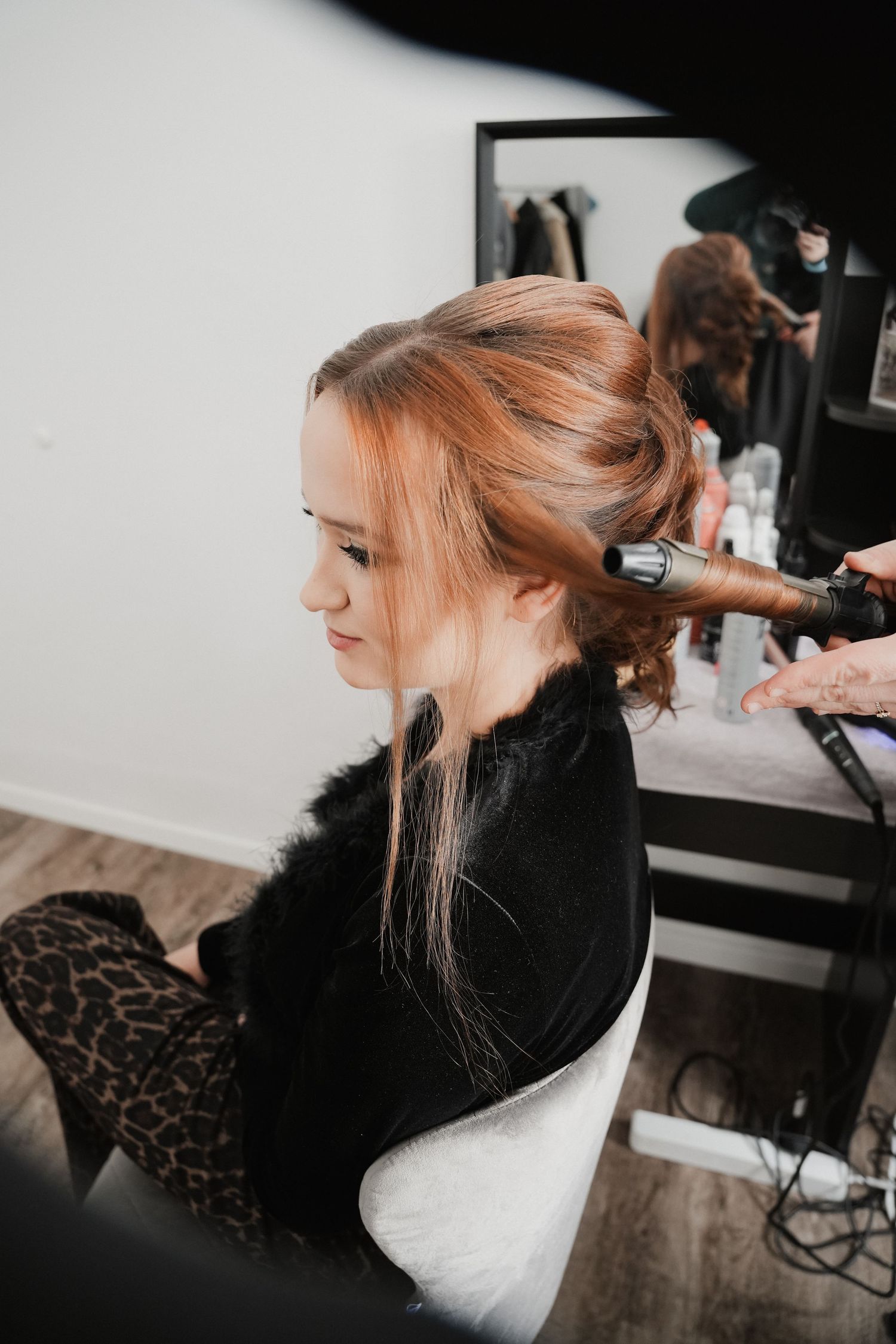 A hairstylist curling a young woman's auburn updo in a modern salon, reflected in a mirror.