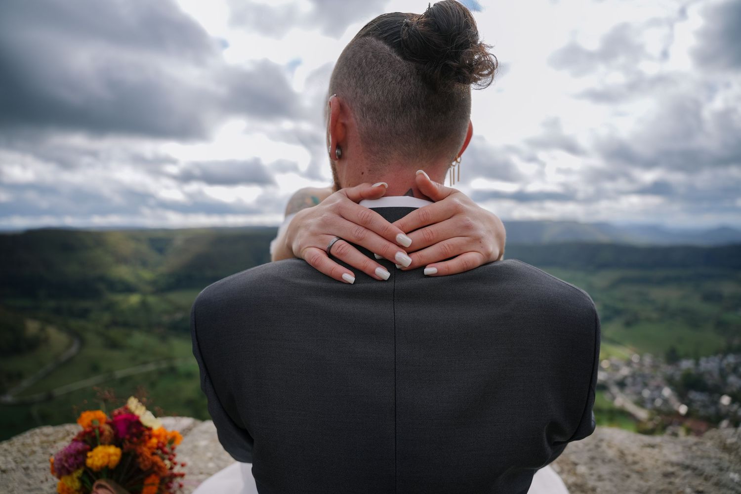 Bride's hands rest on a groom's shoulders as they overlook a scenic green valley under cloudy skies, with a bouquet in the corner.