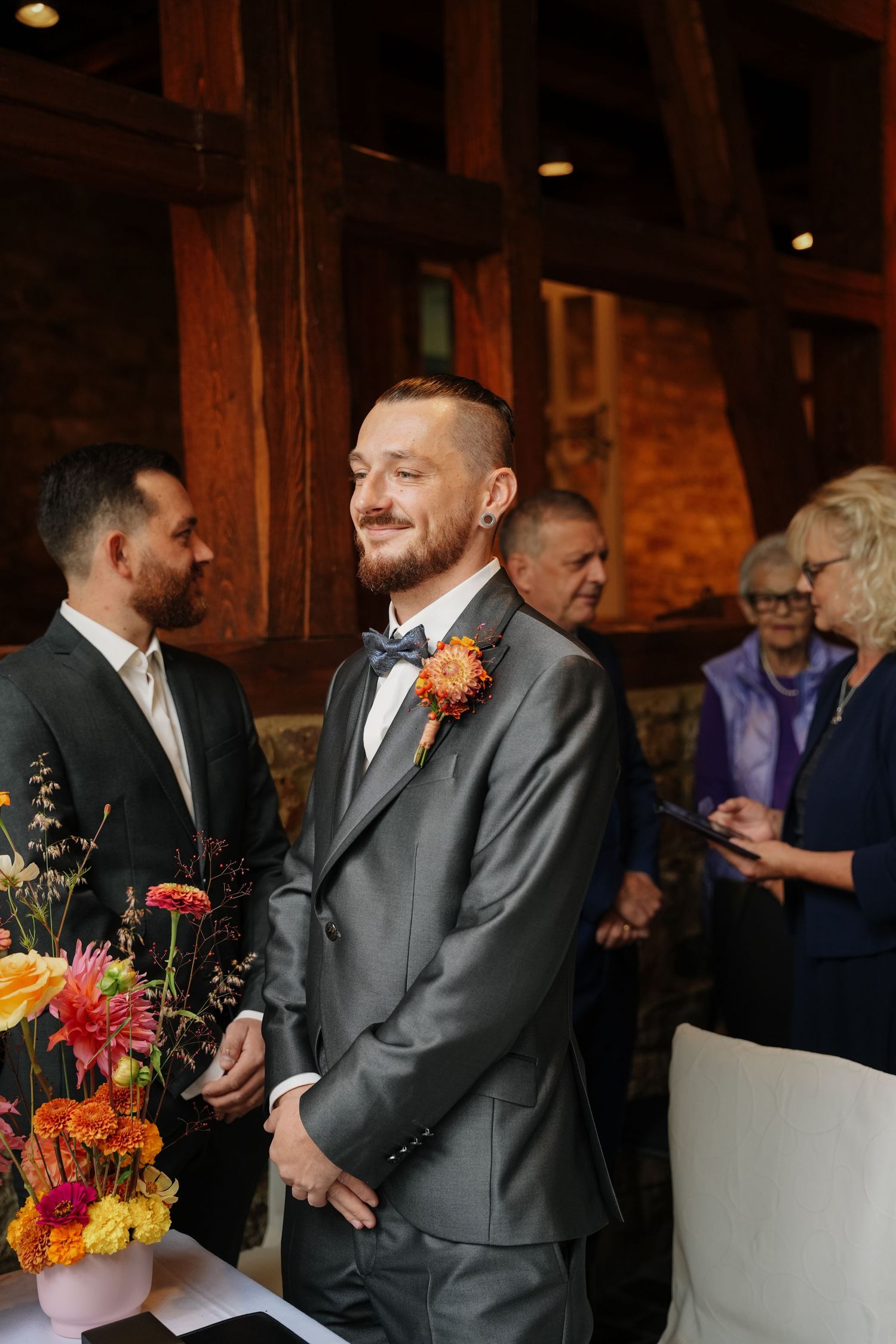 Smiling groom in a gray suit with blue bow tie and orange floral boutonniere stands at a rustic wedding ceremony beside colorful flowers.