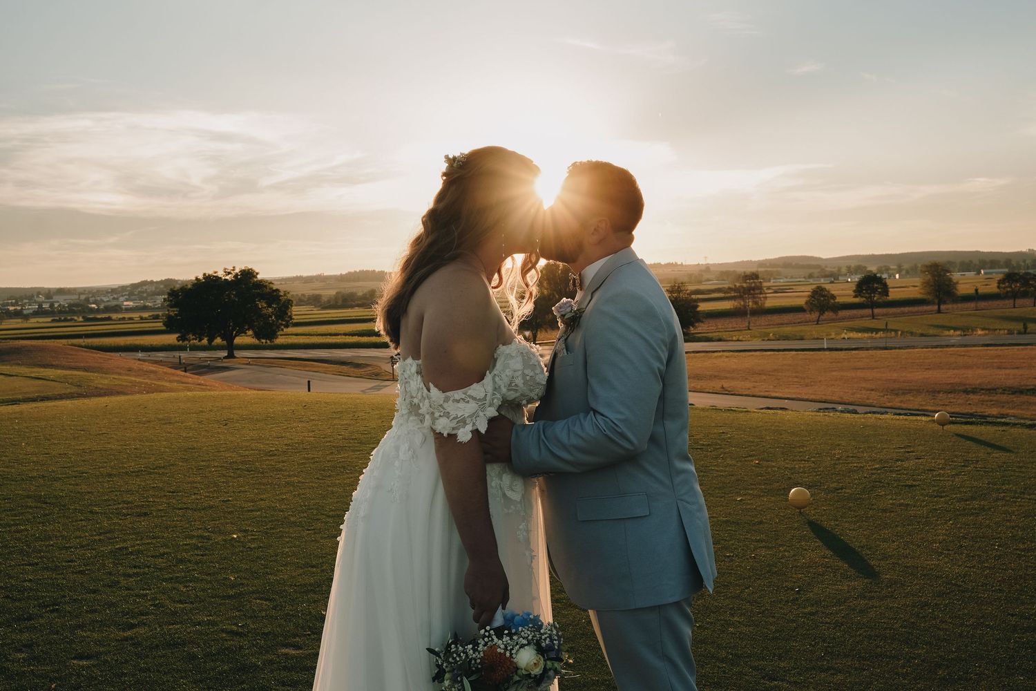Bride and groom sharing a golden sunset kiss on a lush green golf course, with rolling hills in the background.
