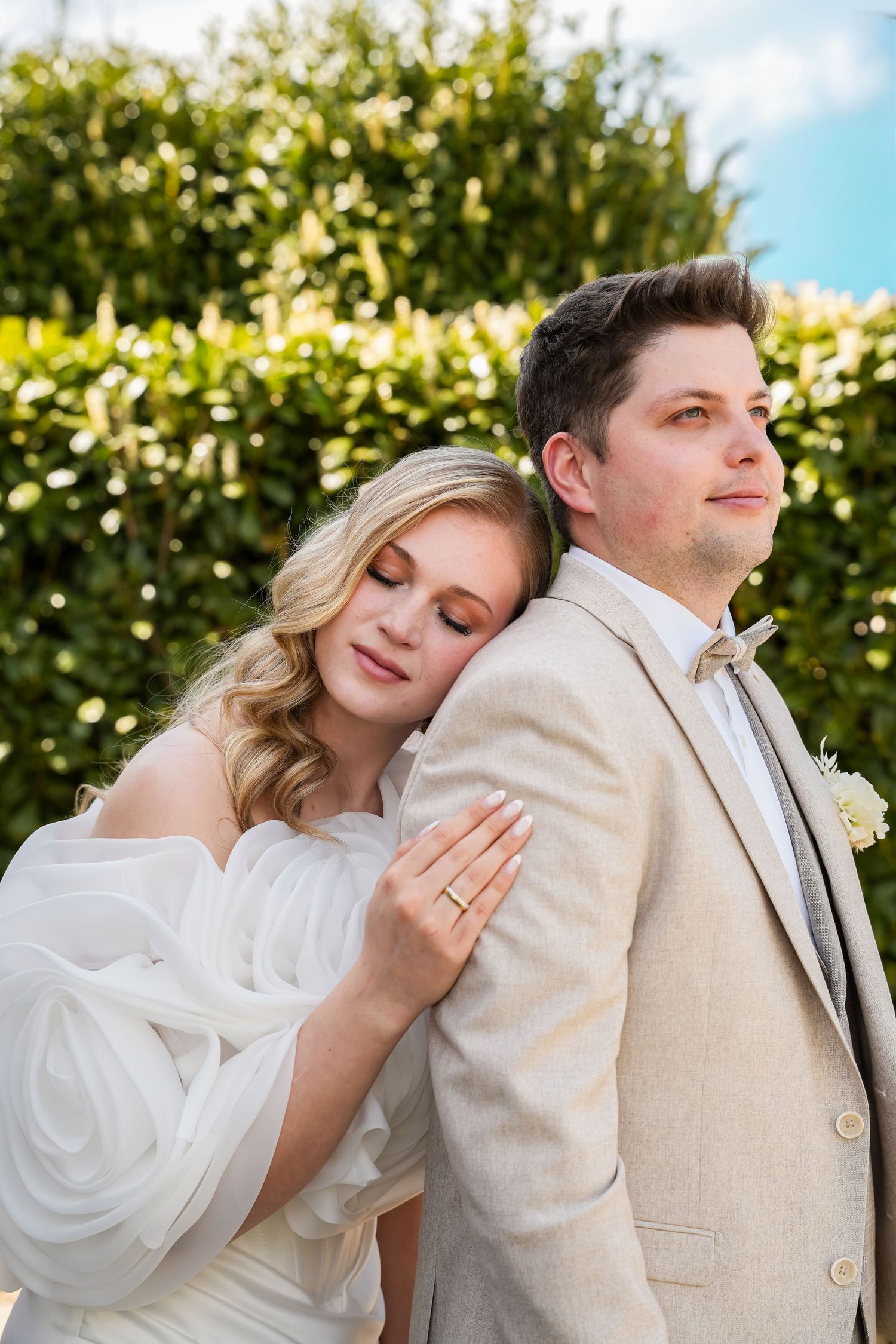 Bride in white dress leans on groom in beige suit with bow tie, posing together in front of lush green hedges.