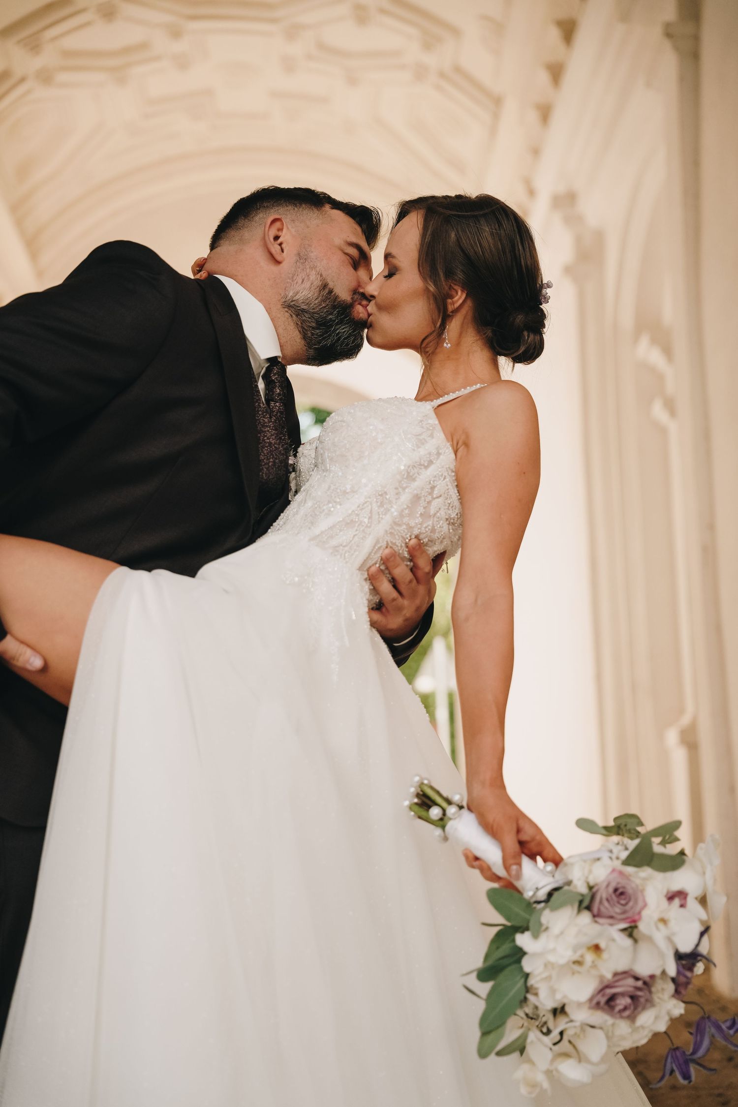 Bride and groom sharing romantic kiss under ornate arch, bride holding white and lavender floral bouquet.